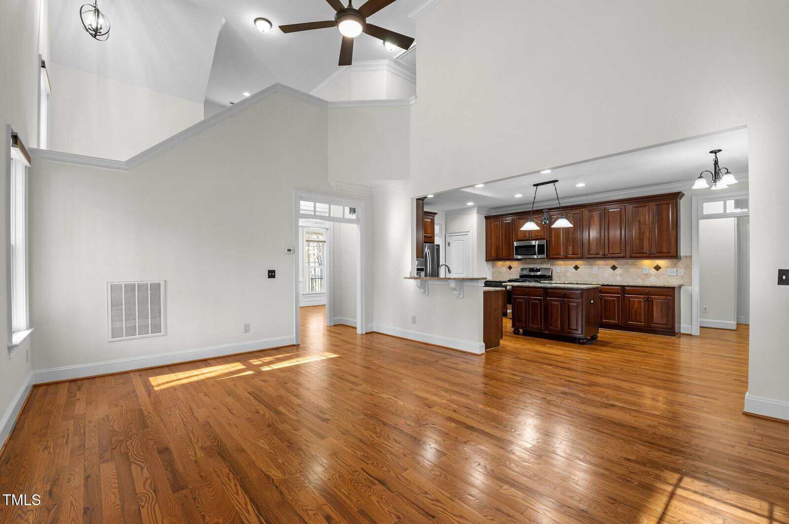 A spacious, well-lit room with wooden flooring connects to an open kitchen featuring dark wood cabinets, stainless steel appliances, and a light granite countertop. Recessed and pendant lighting adorn the ceiling. Natural light filters in from large windows.