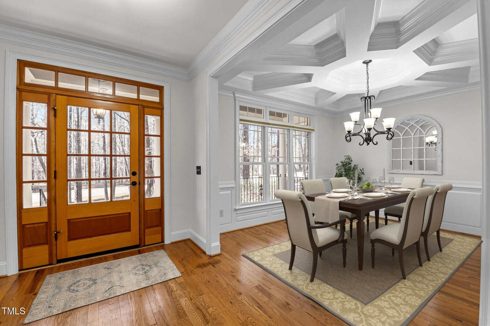 Dining room with a wooden floor and coffered ceiling. A table with six chairs, set with white dishes and green decor, sits under a chandelier. Large windows and a glass-paneled wooden door provide natural light. A patterned rug is near the entrance.