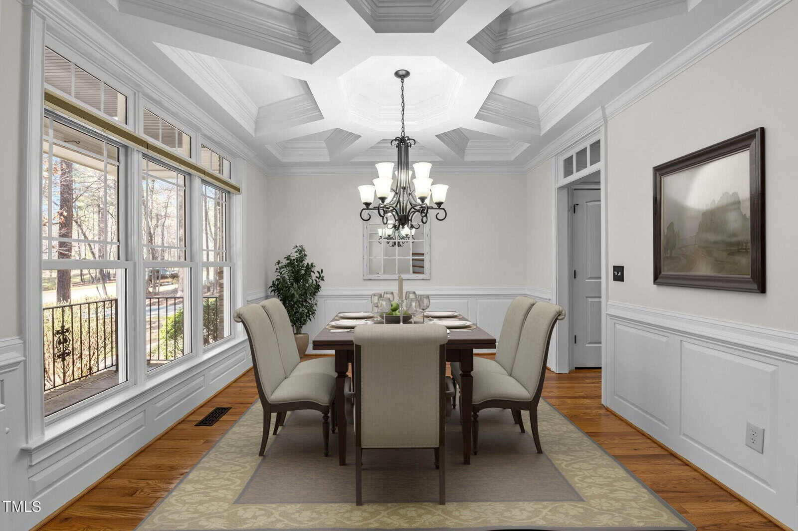 A dining room with a rectangular table and six cushioned chairs. A chandelier hangs above the table. Large windows on the left let in natural light. The walls are light-colored with wainscoting, and there is a framed picture on one wall.