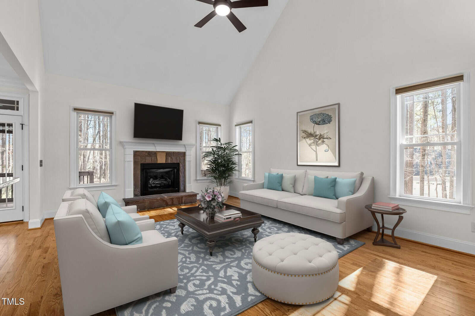 Spacious living room with a vaulted ceiling, featuring a white couch and chairs with light blue cushions, a large rug, a fireplace beneath a wall-mounted TV, and windows providing natural light. There's a plant in the corner and a round ottoman.