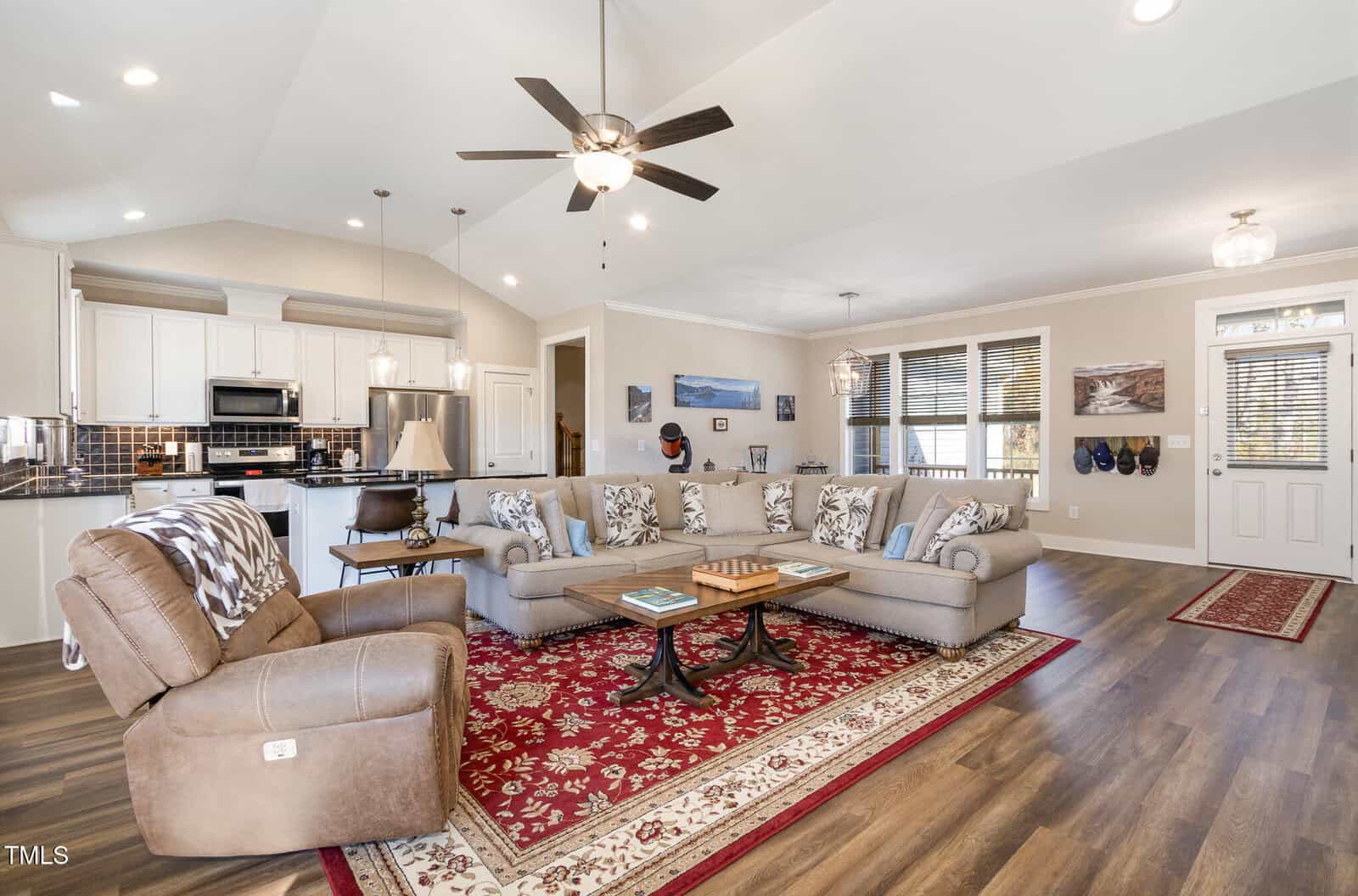 A spacious living room with an open kitchen layout. The room has a large beige sectional sofa, a brown recliner, a patterned red rug, and a ceiling fan. The kitchen area has white cabinets and stainless steel appliances. Wall art and decor are visible.