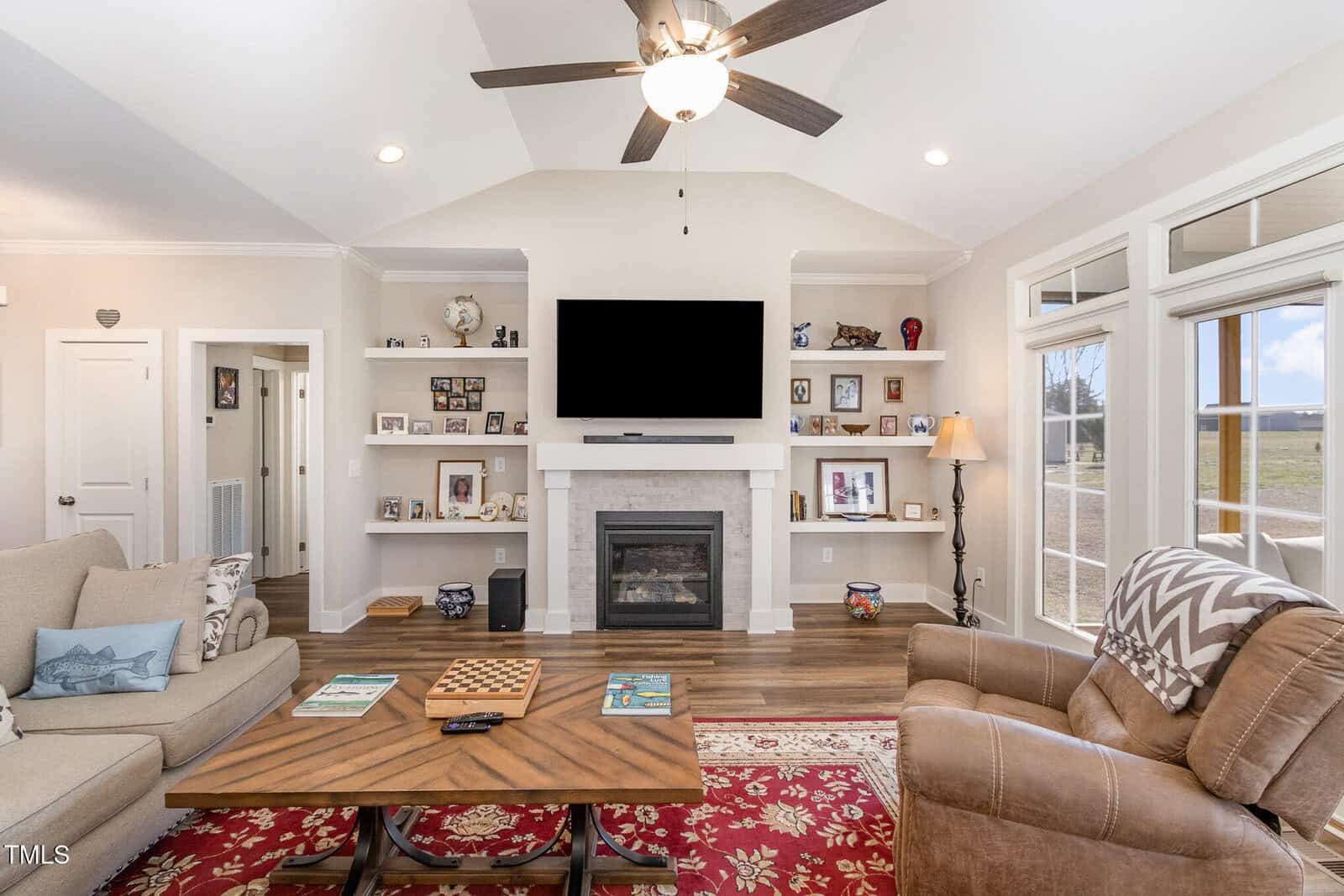 A living room with a beige sofa and brown armchair, centered around a wooden coffee table. A fireplace with a wall-mounted TV is flanked by shelves with photos and decor. A ceiling fan hangs above. Windows on the right allow natural light in.