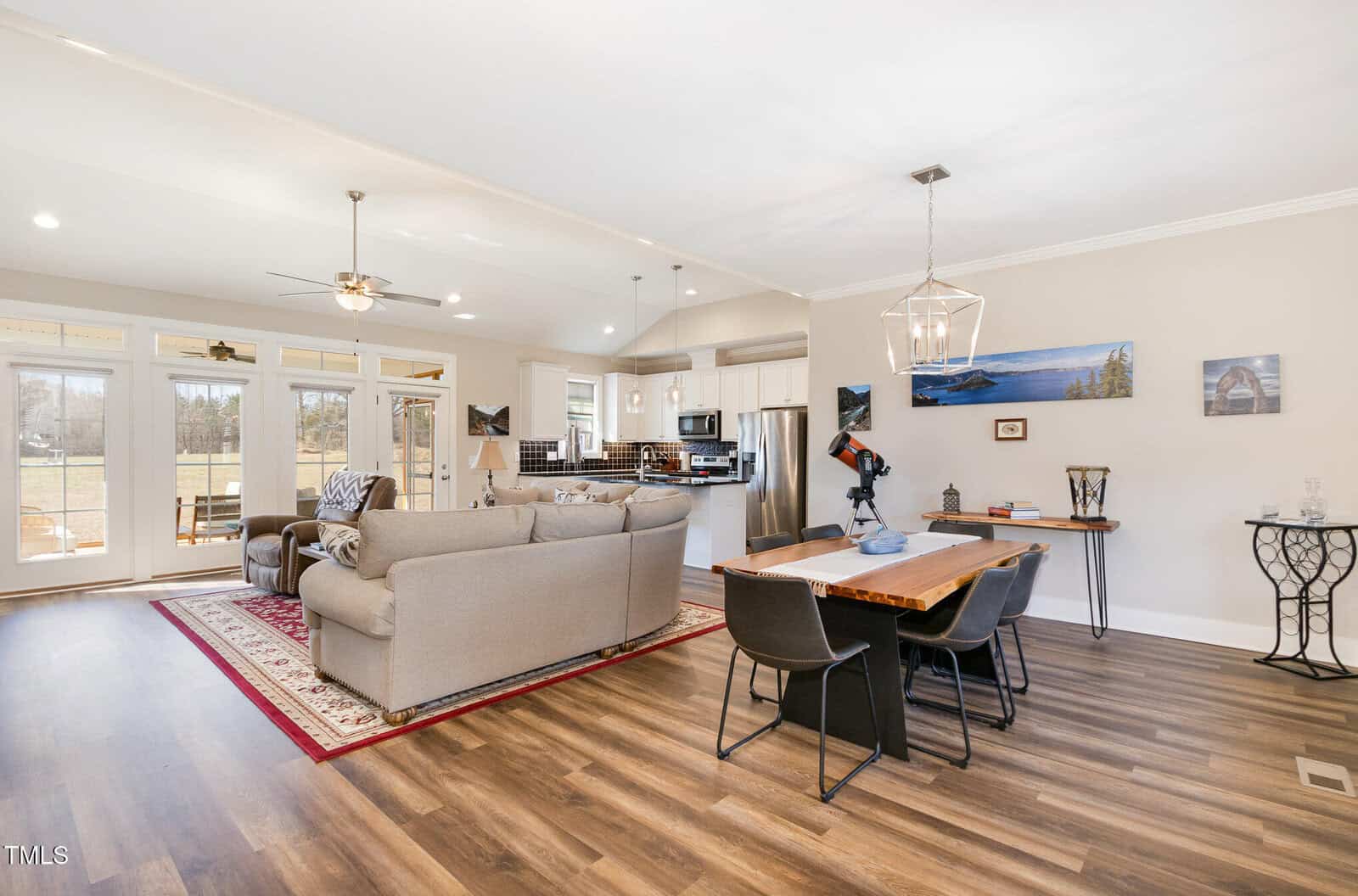 Open-concept living and dining area with large windows. Features a gray sectional sofa, armchair, area rug, and ceiling fan. Dining area has a wooden table with black chairs and a modern light fixture. Brown hardwood floors throughout.