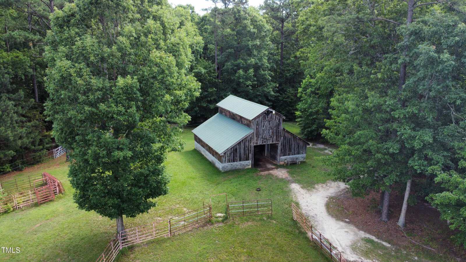 A wooden barn with a green metal roof sits in a grassy, fenced area surrounded by tall trees. A dirt path leads up to the barn, and metal gates are visible near the entrance.
