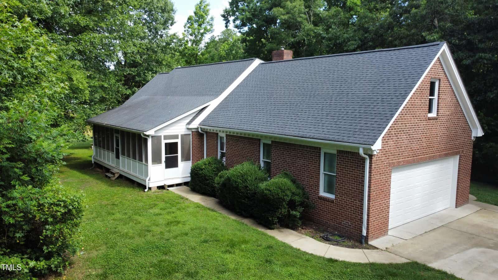 A single-story brick house with a dark gray shingled roof, a white two-car garage, and a screened-in porch. The house is surrounded by green grass, bushes, and trees.