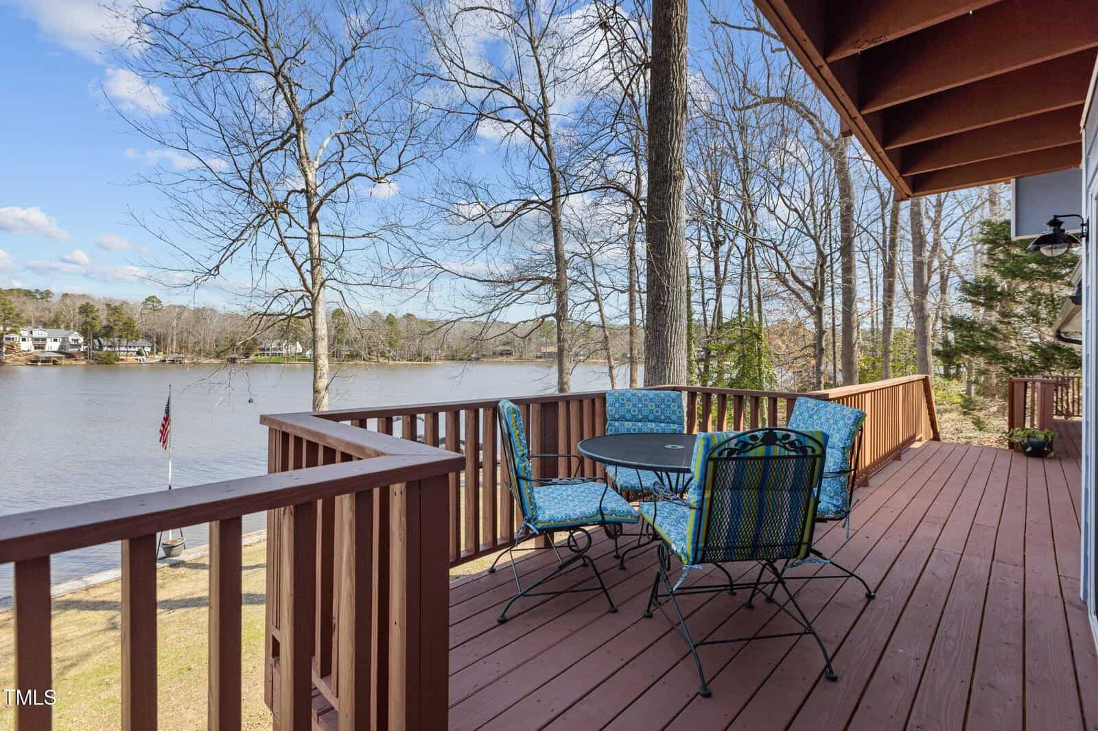 A wooden deck with a round table and four blue cushioned chairs overlooks a lake. Trees surround the area, some without leaves. An American flag is visible near the water, and houses are seen across the lake.
