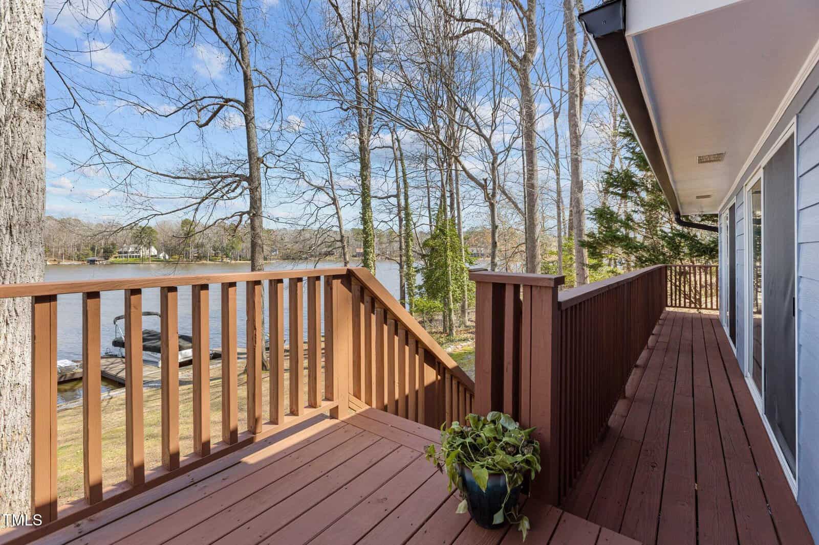 A wooden deck with a railing overlooks a lake and trees. A potted plant sits in the corner of the deck. The house exterior is visible on the right, and the sky is partly cloudy.