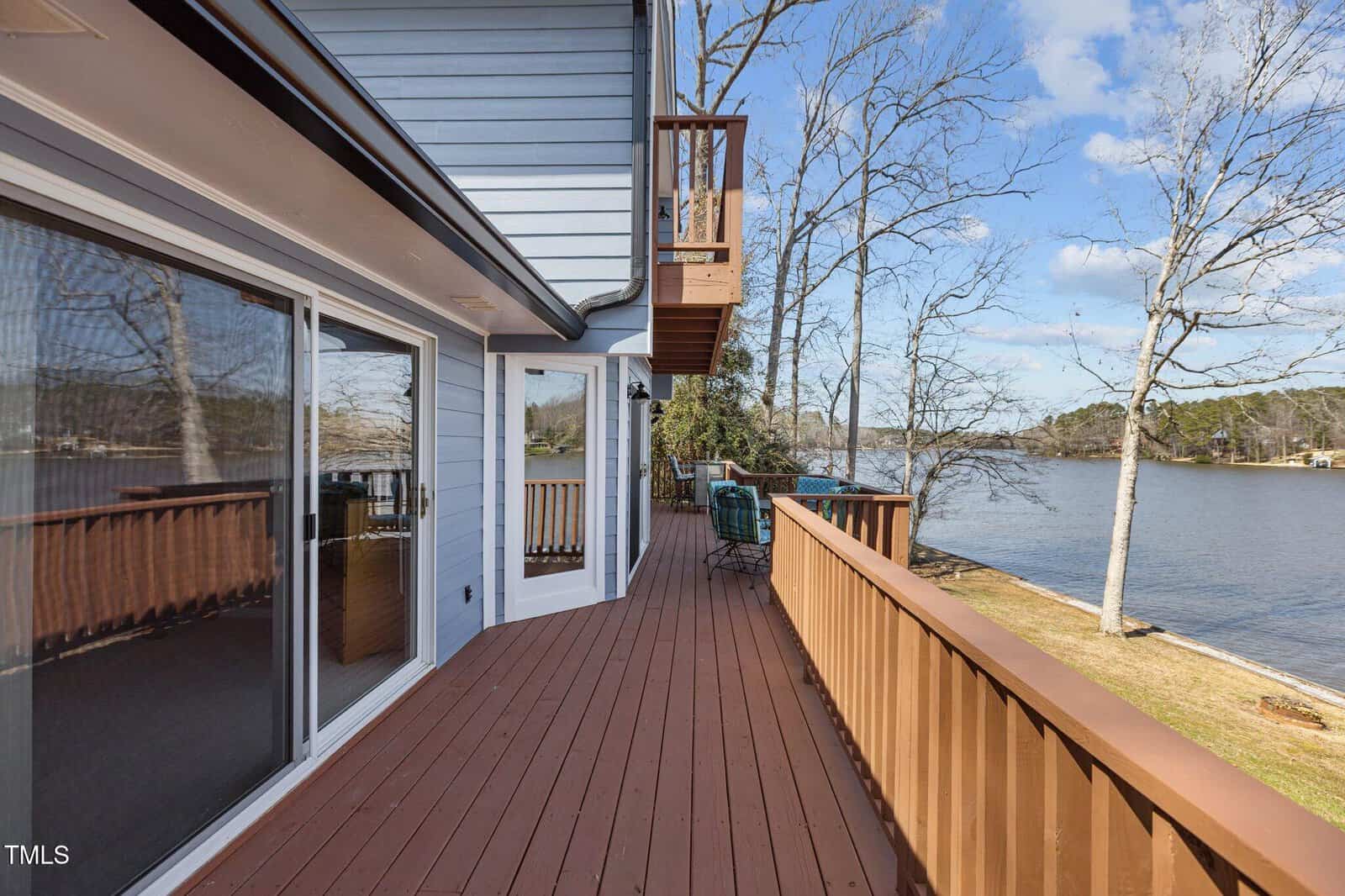 A wooden deck wraps around the side of a light blue house overlooking a lake. The deck includes a railing, outdoor seating, and is bordered by leafless trees. Blue sky and scattered clouds are visible.
