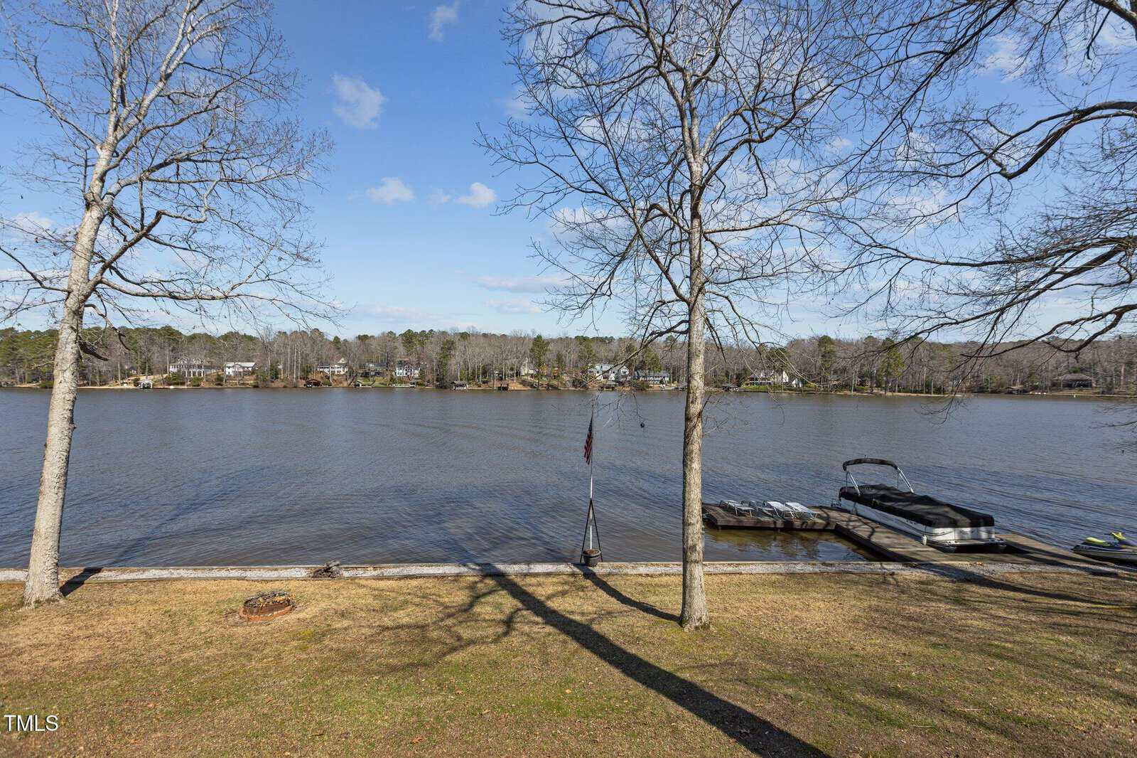 View of a calm lake bordered by trees, with a wooden dock extending into the water. A pontoon boat and jet ski are moored at the dock. The sky is mostly clear, and leafless trees stand in the foreground on a grassy shore.