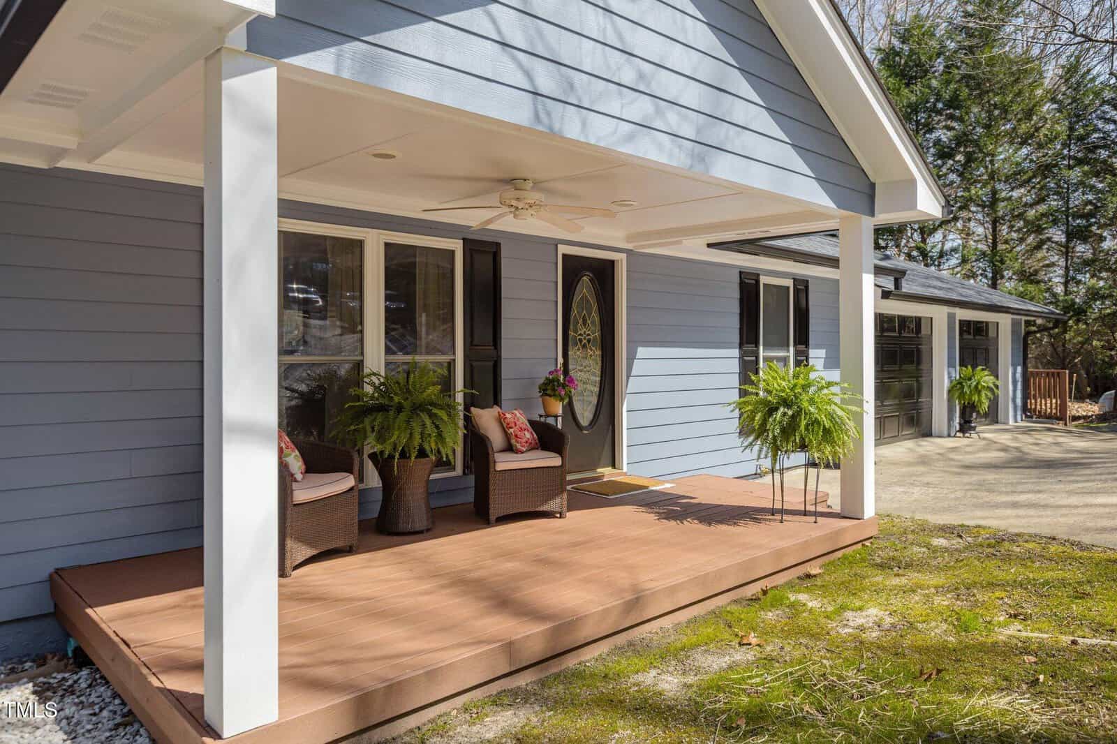 A blue house with white trim features a covered front porch with two chairs, two potted ferns, and a ceiling fan. A two-car garage is attached on the right side. The porch overlooks a grassy yard.