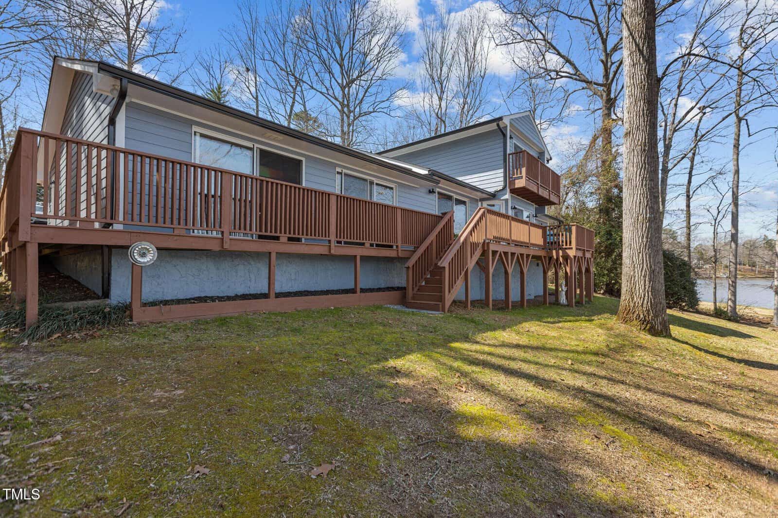 A light blue house with a wooden deck and stairs sits on a grassy yard. Leafless trees surround the property, and a body of water is visible in the background under a partly cloudy sky.