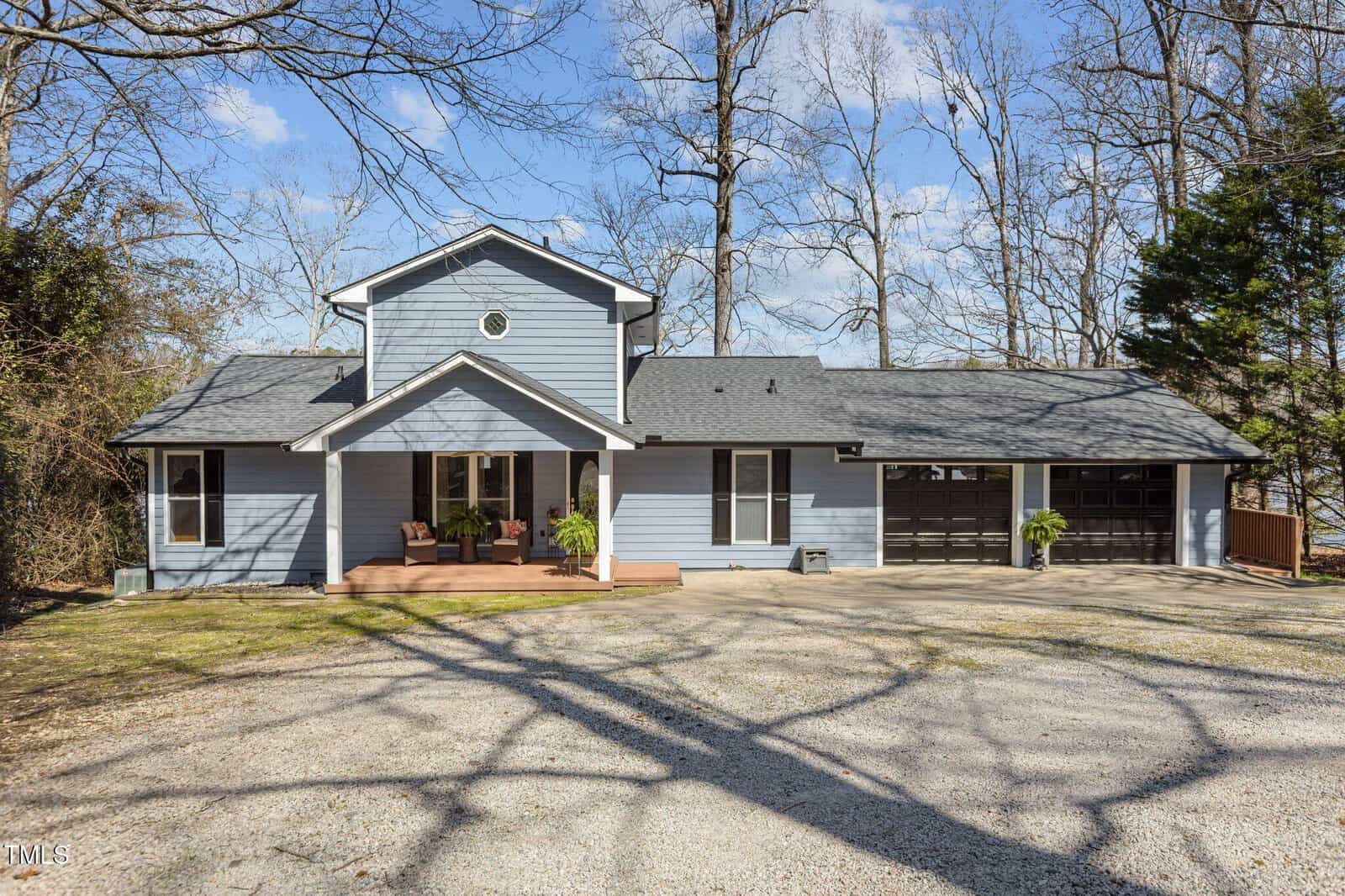 A two-story blue house with white trim, a covered front porch, and a gravel driveway. There are leafless trees around the property and potted plants by the garage. The sky is partly cloudy.