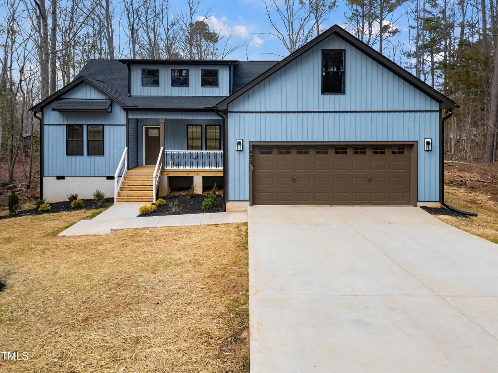 A modern, two-story house with blue siding, white trim, a brown double garage door, front porch with stairs, and a freshly paved driveway. The yard is partially landscaped with small plants. Trees are visible in the background.