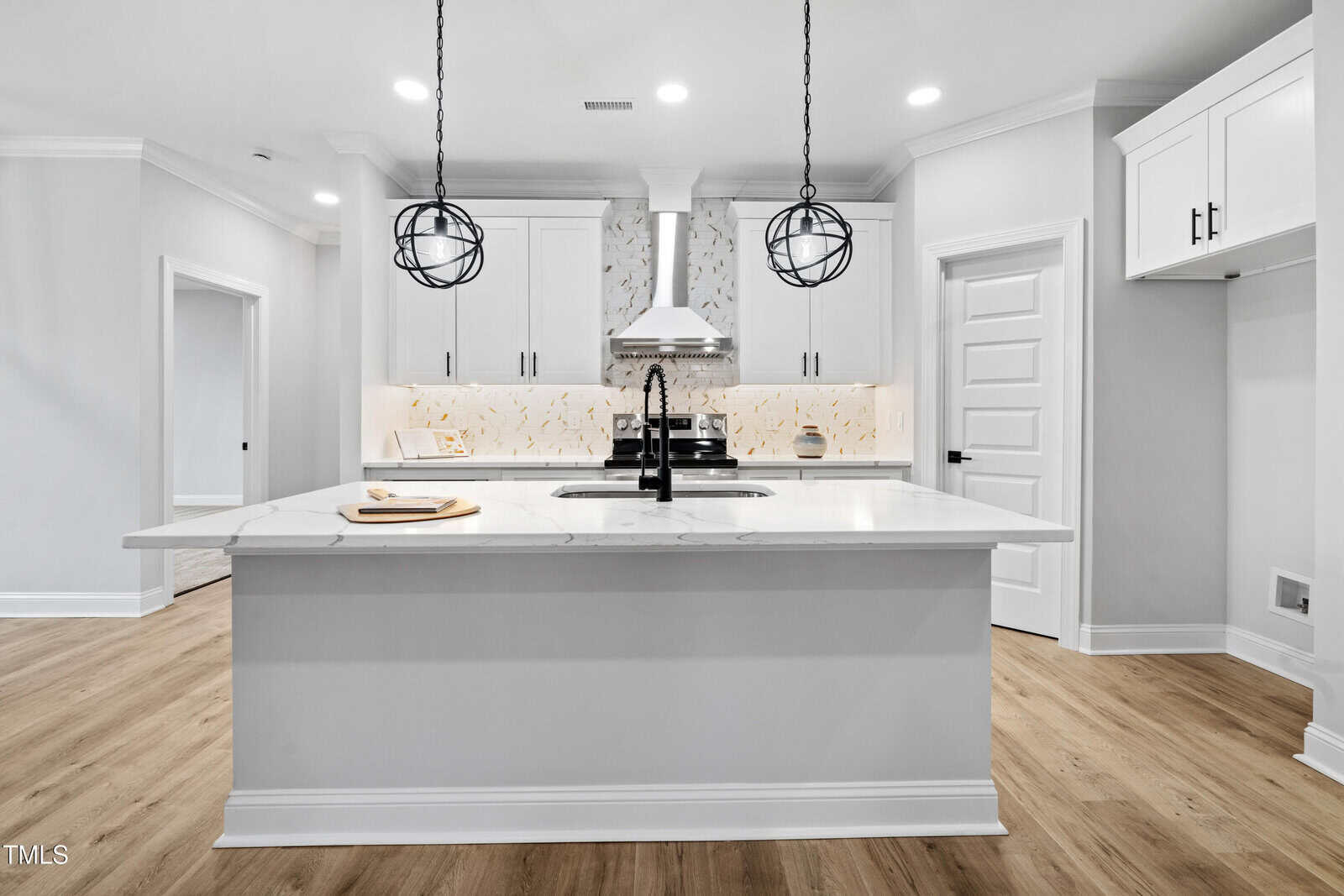 Modern kitchen with white cabinets, marble island with sink, light wood floors, two spherical pendant lights, white backsplash, stainless steel stove with hood, and three white doors in the background.