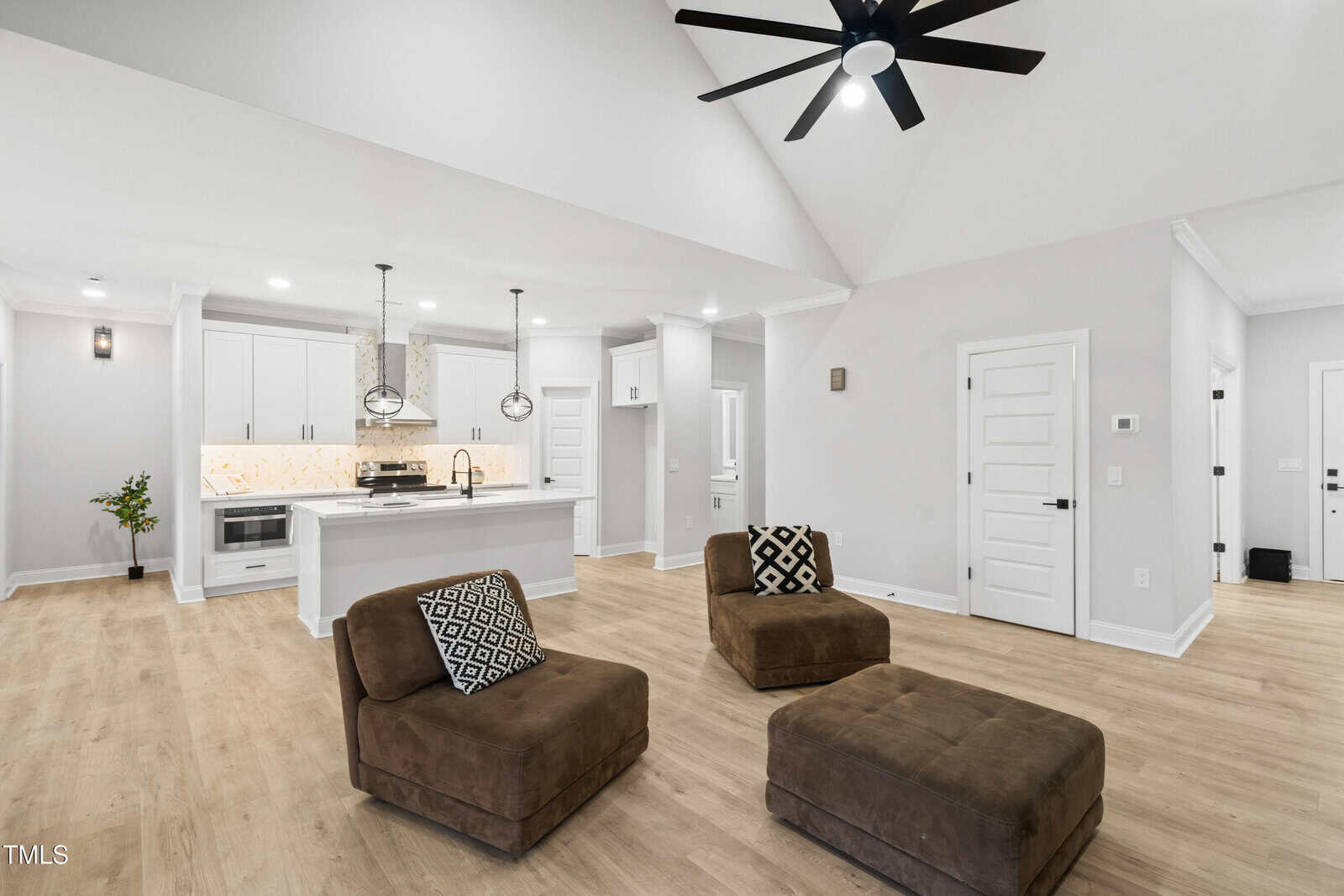 Modern open-concept living room and kitchen with white cabinets, a large island, pendant lights, tan wood floors, two brown chairs with patterned pillows, and a matching ottoman. Ceiling fan and recessed lighting are visible.