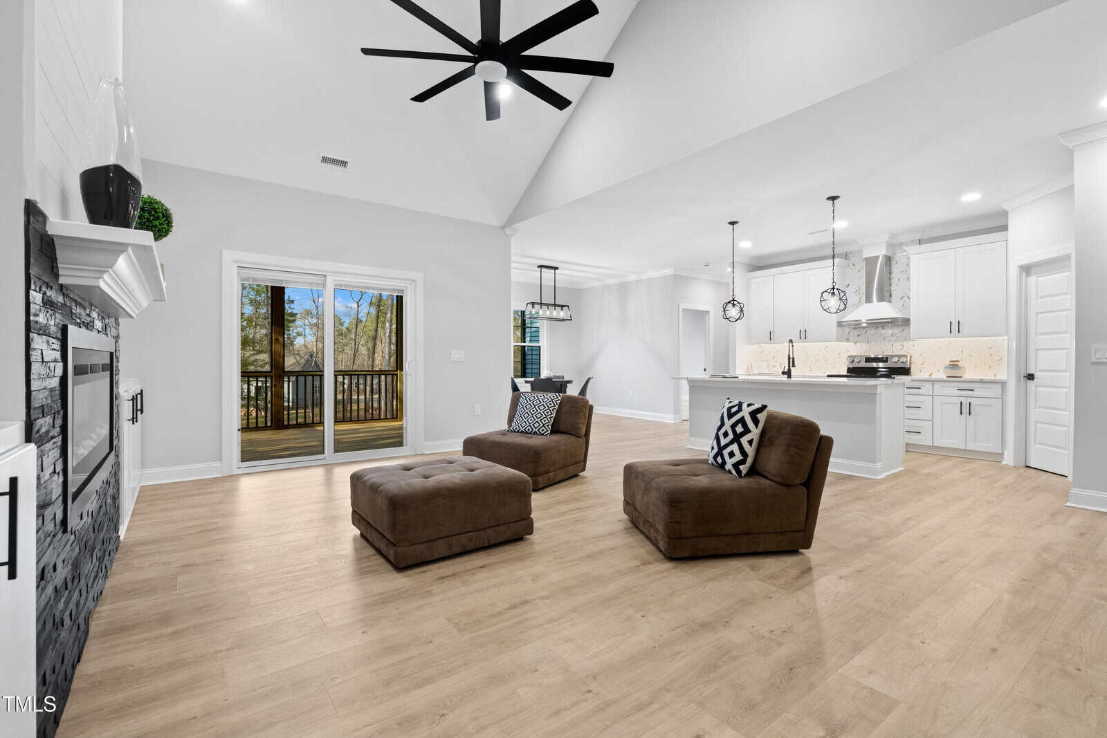 Open-concept living room and kitchen with light wood floors, two brown chairs with matching ottomans, modern ceiling fan, fireplace, and sliding doors leading to a deck with a wooded view.