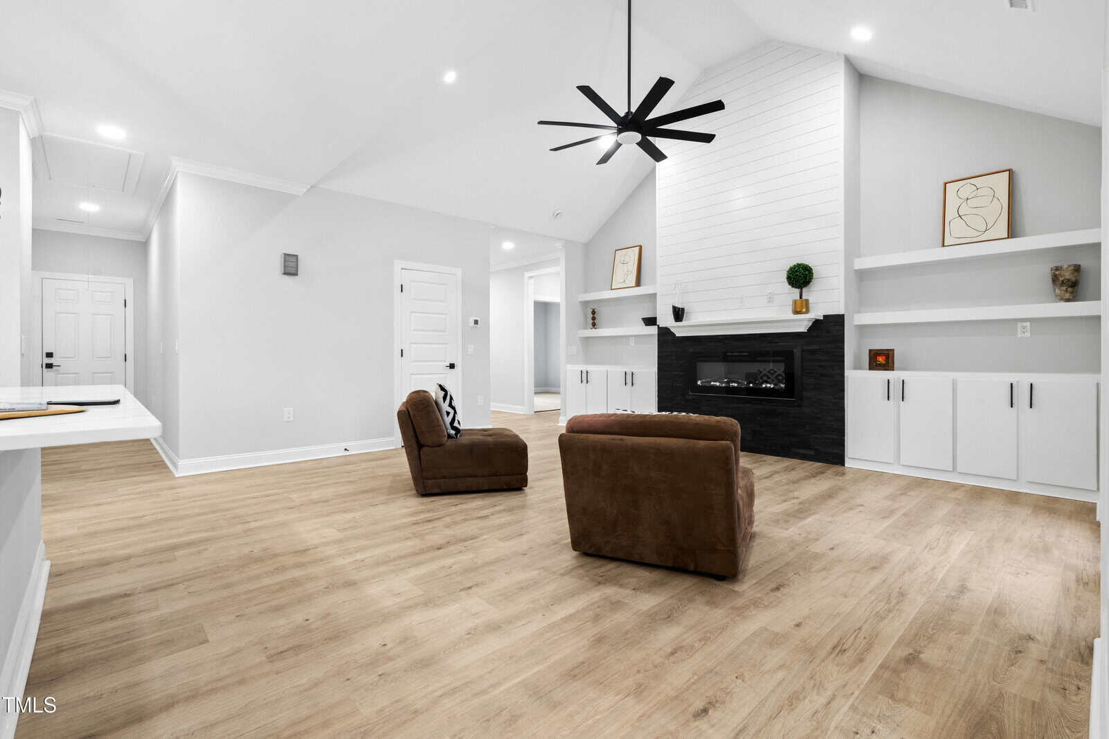 Modern living room with light wood floors, two brown chairs, a black fireplace, white built-in cabinets with shelves, and a geometric black ceiling fan. Walls are white, and decor is minimal. A hallway and entry door are visible.