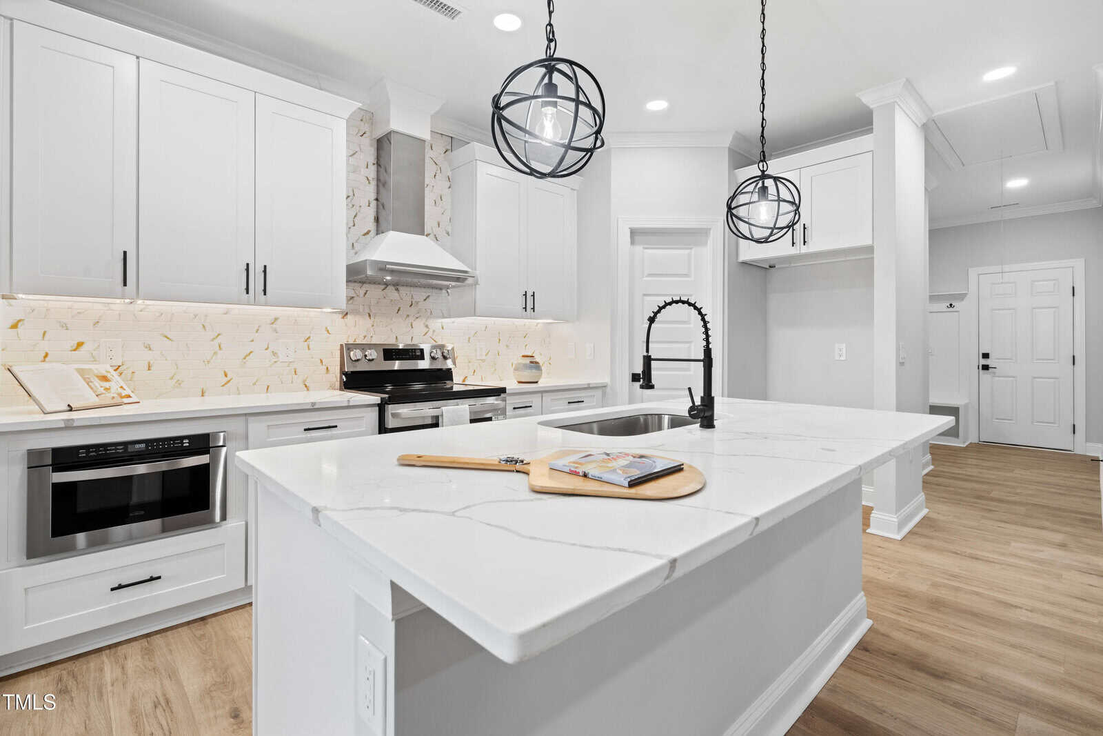 Modern kitchen with white cabinets, stainless steel appliances, marble countertops, a central island with a black faucet, two pendant lights, and light wood flooring. A book and cutting board are on the island.