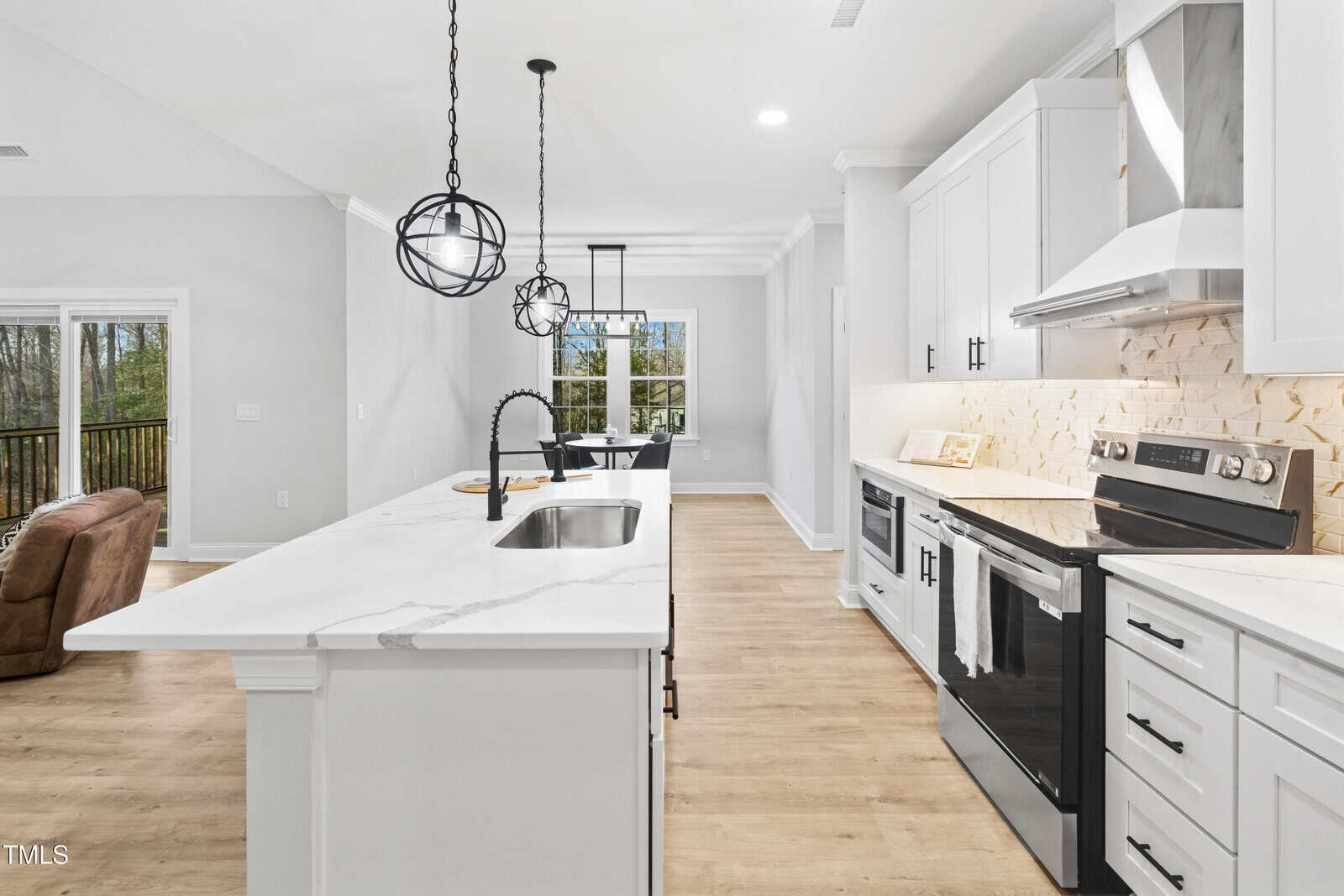 A modern kitchen with white cabinets, a marble island with a sink, stainless steel appliances, two pendant lights, light wood flooring, and a view of the dining area and sliding glass doors to a deck.