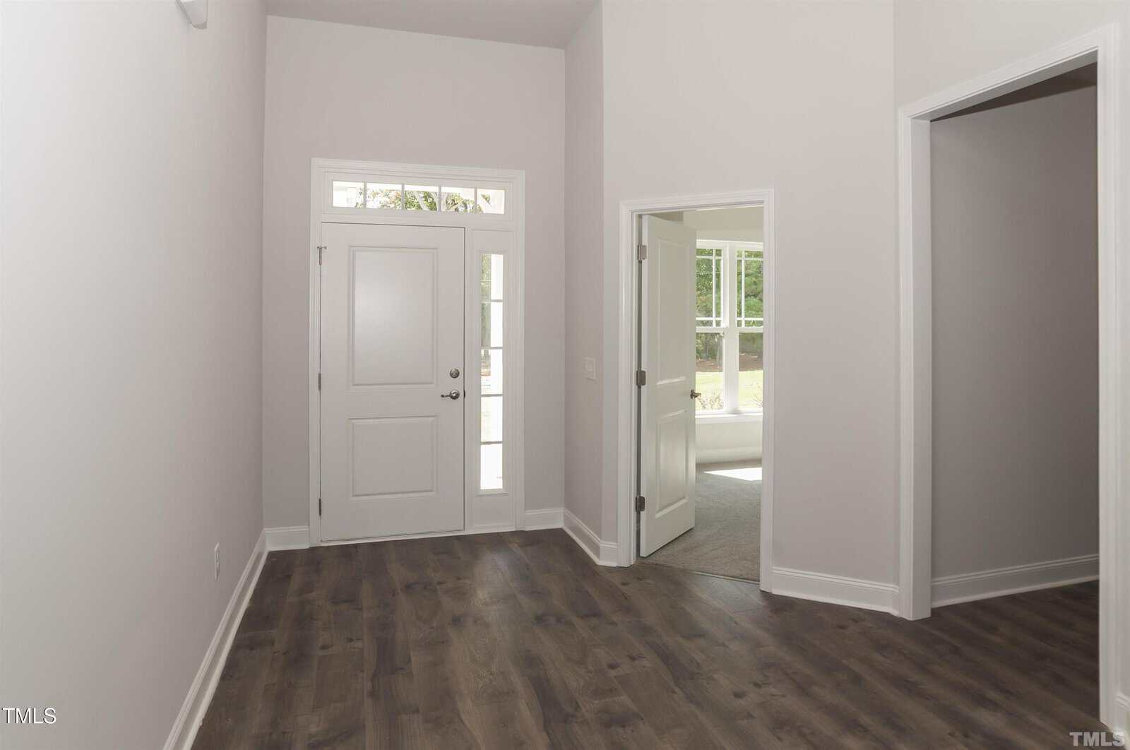 A hallway with dark wood flooring, white walls, and a white front door with window panels. An open doorway on the right leads to a bright room with carpet and large windows.
