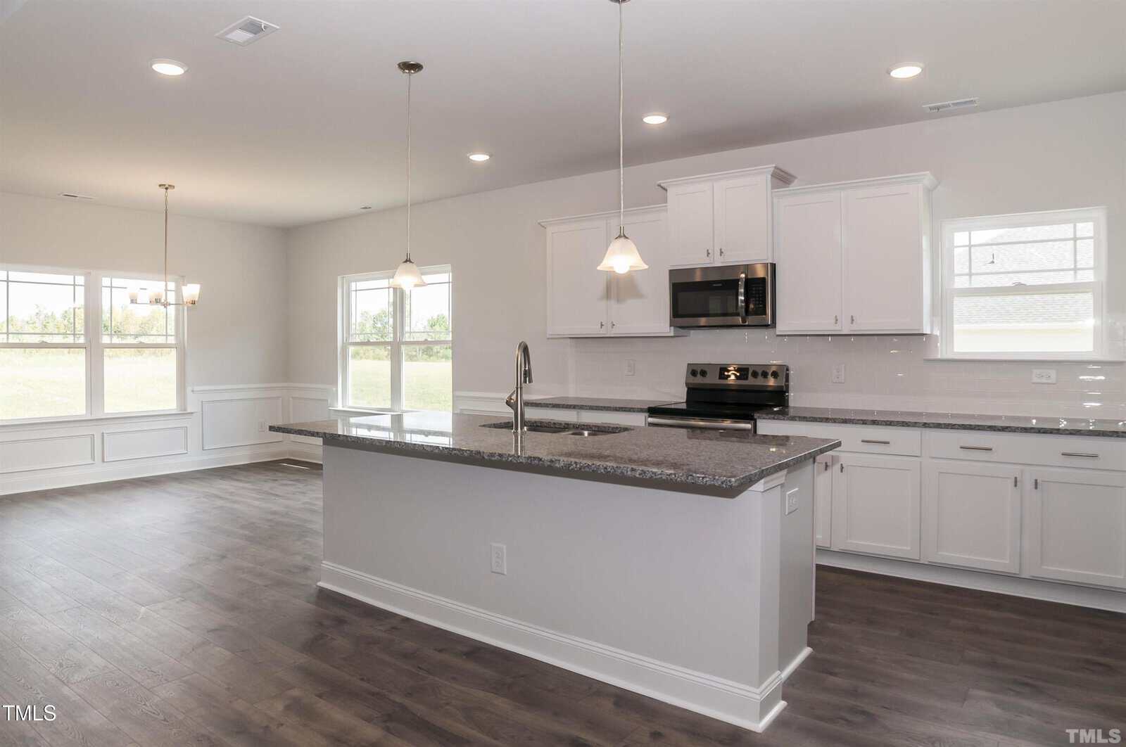 A modern kitchen with white cabinets, stainless steel appliances, a granite island with a sink, pendant lights above the island, and an adjacent dining area with large windows letting in natural light.