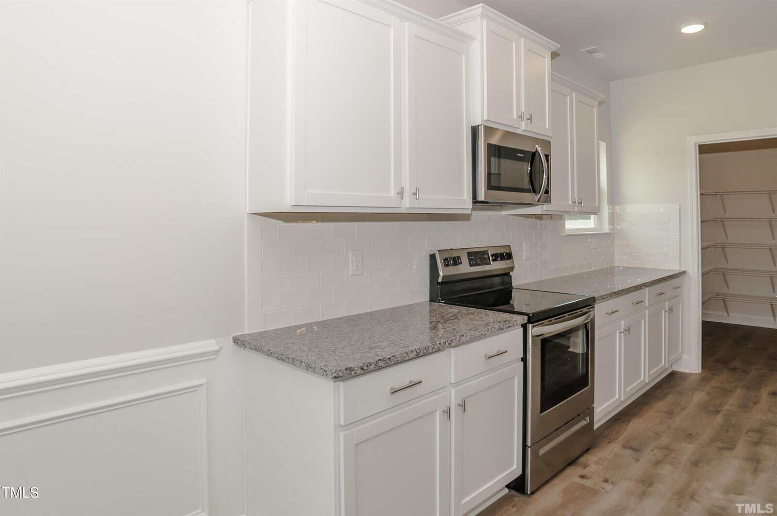 Modern kitchen with white cabinets, stainless steel oven and microwave, granite countertops, white subway tile backsplash, and wood-look flooring. There is an open doorway leading to a pantry with shelving.