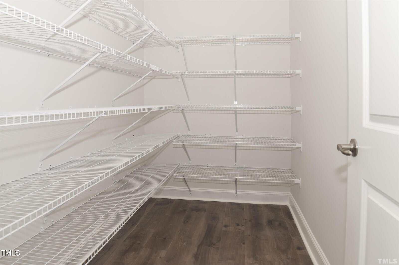 A walk-in pantry with white wire shelving on three walls and a dark wood floor. The walls are light-colored, and the pantry door is white with a silver handle.