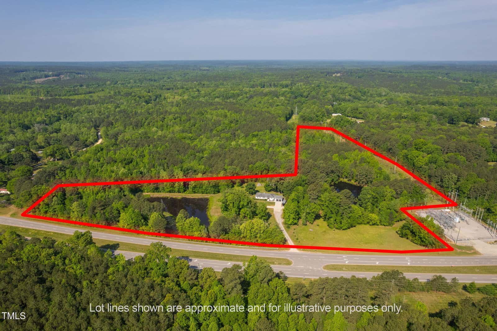Aerial view of a large, wooded property outlined in red, bordered by a highway in the foreground. The land includes a pond, a few buildings, open grassy areas, and is surrounded by dense forest. Text notes lot lines are approximate.