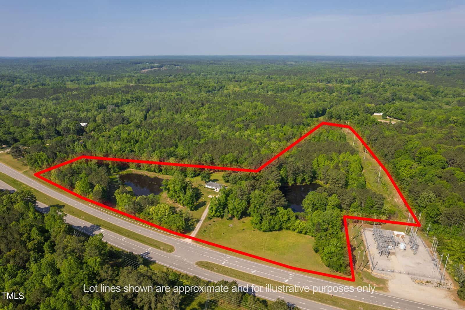 Aerial view of a wooded property outlined in red, bordered by a highway on one side and an electrical substation on another, with a small pond and several trees within the lot. Lot lines shown are approximate.