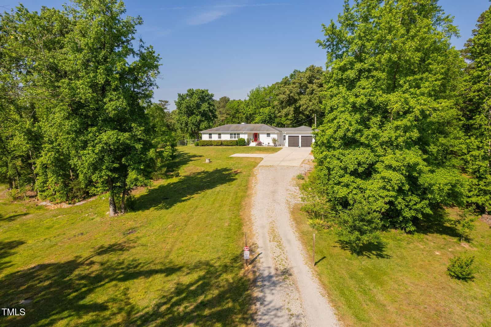 A long gravel driveway leads to a single-story white house with an attached garage, surrounded by green grass and tall trees under a clear blue sky.
