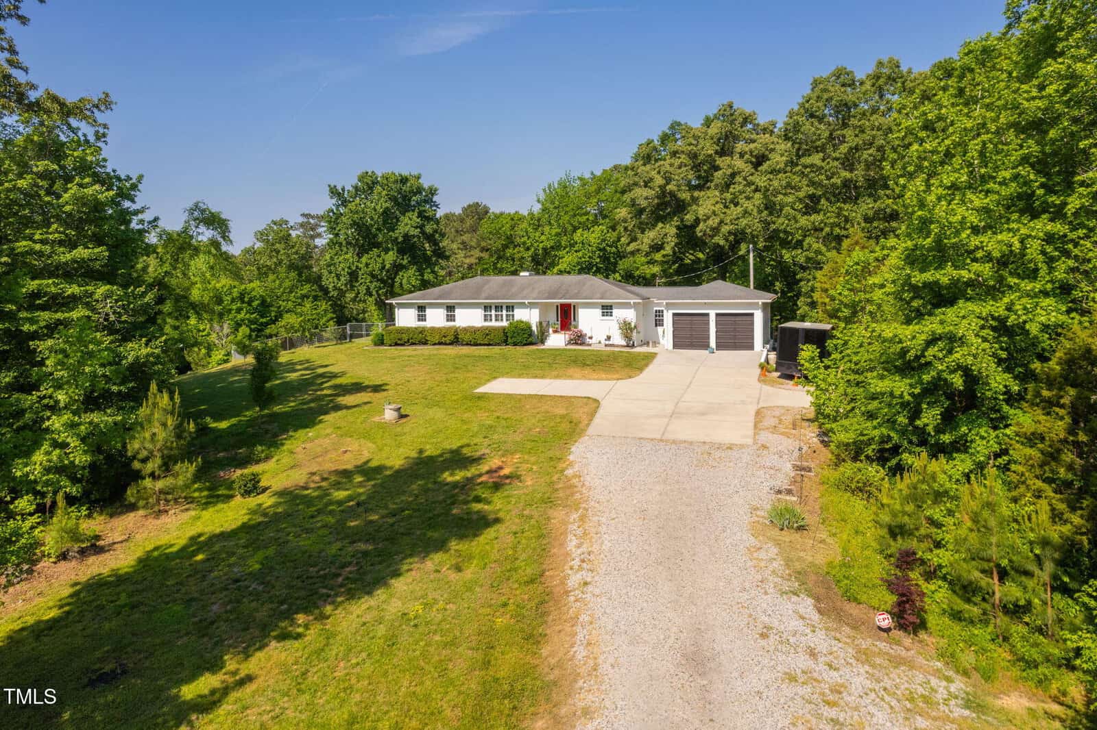 A single-story white house with a gray roof, red front door, and two-car garage is surrounded by green trees. A gravel driveway leads to a large concrete parking area in front of the house. The lawn is spacious and partially landscaped.