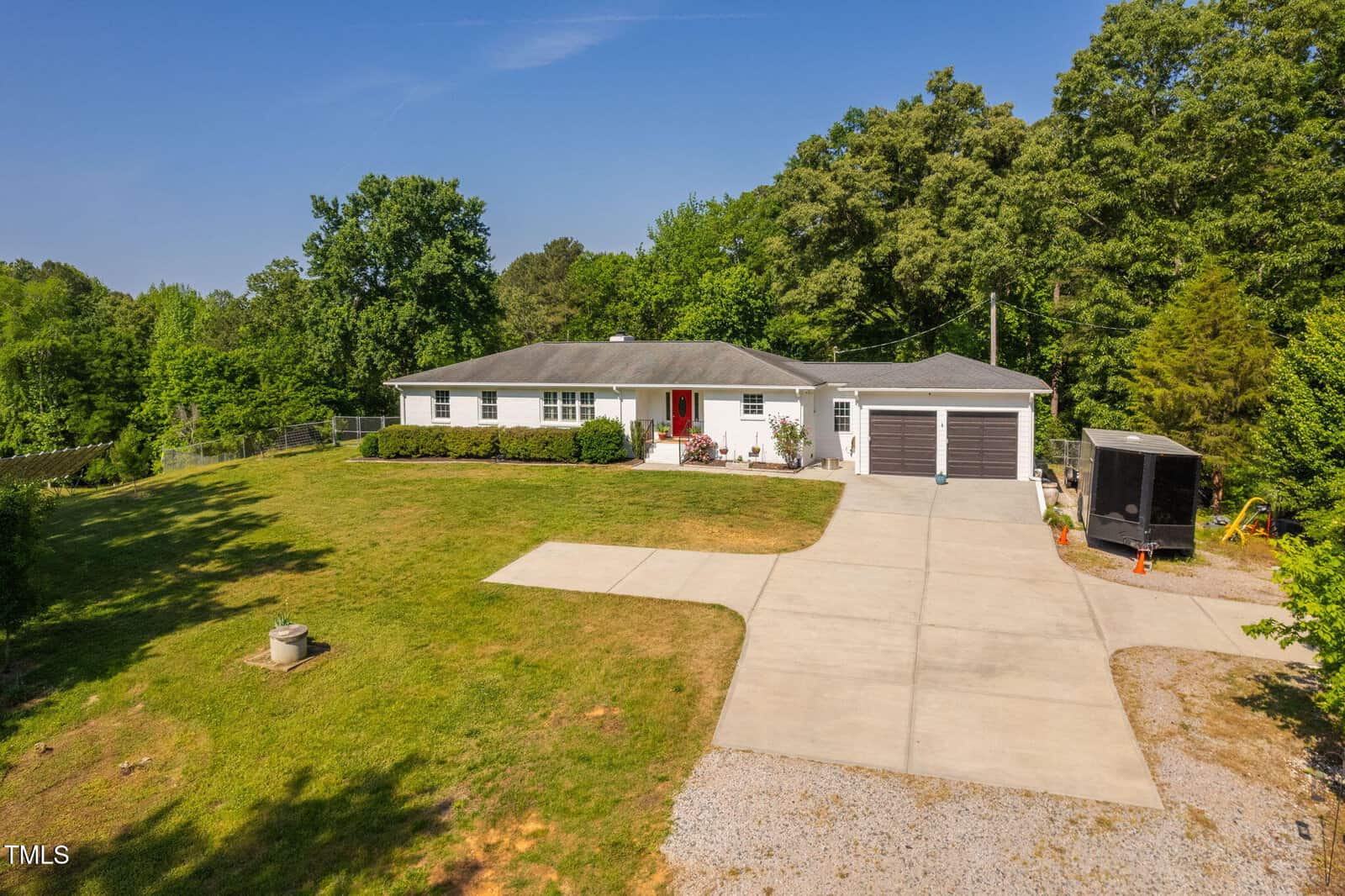 Single-story house with a gray roof, white exterior, and red front door, set on a large grassy yard with a long concrete driveway, two-car garage, and trees in the background. A black trailer is parked beside the house.