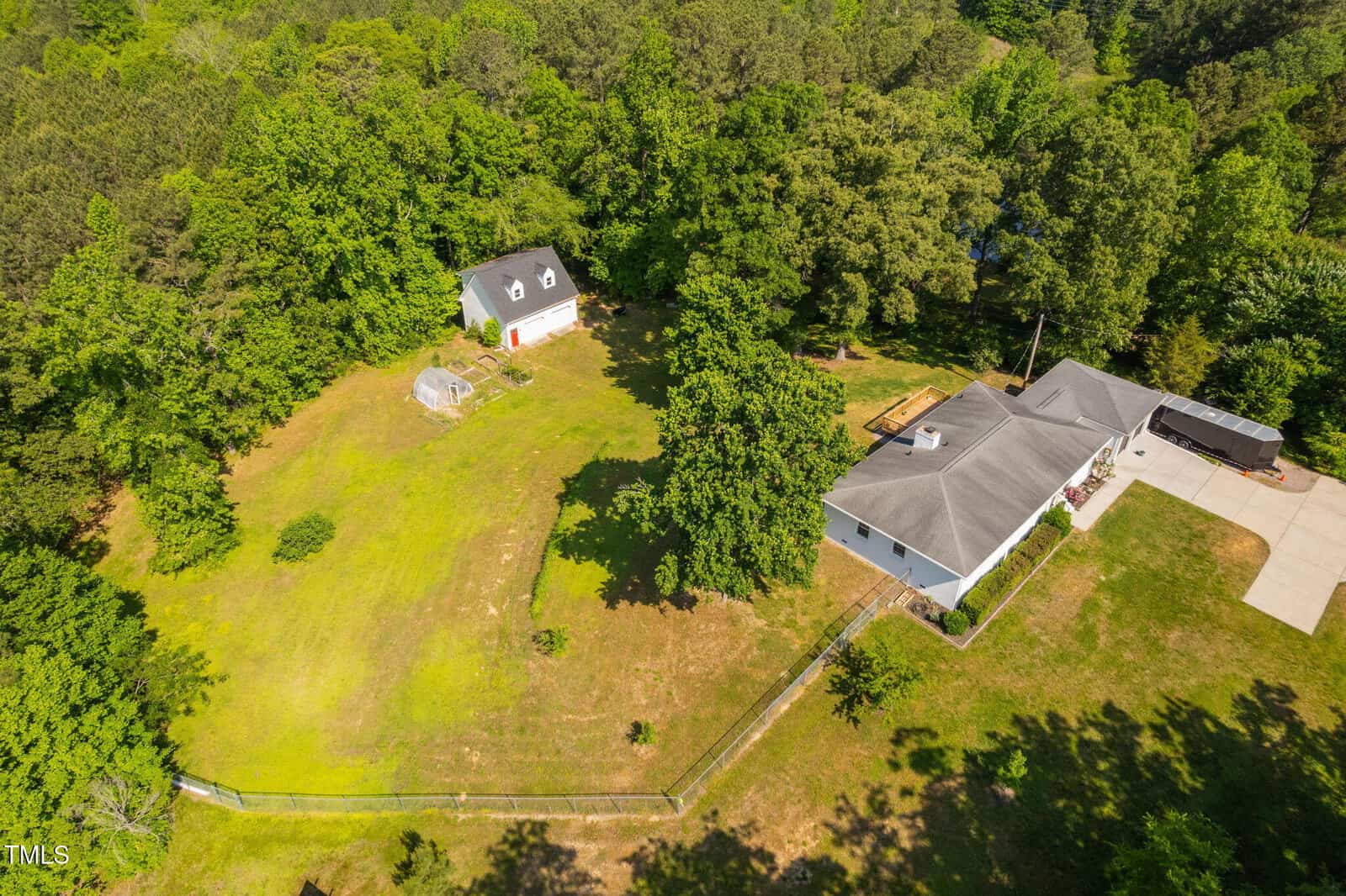 Aerial view of a house with a gray roof, detached garage, fenced yard, small greenhouse, and surrounding trees. A paved driveway leads to the house, which has a deck attached. The property is bordered by dense greenery.