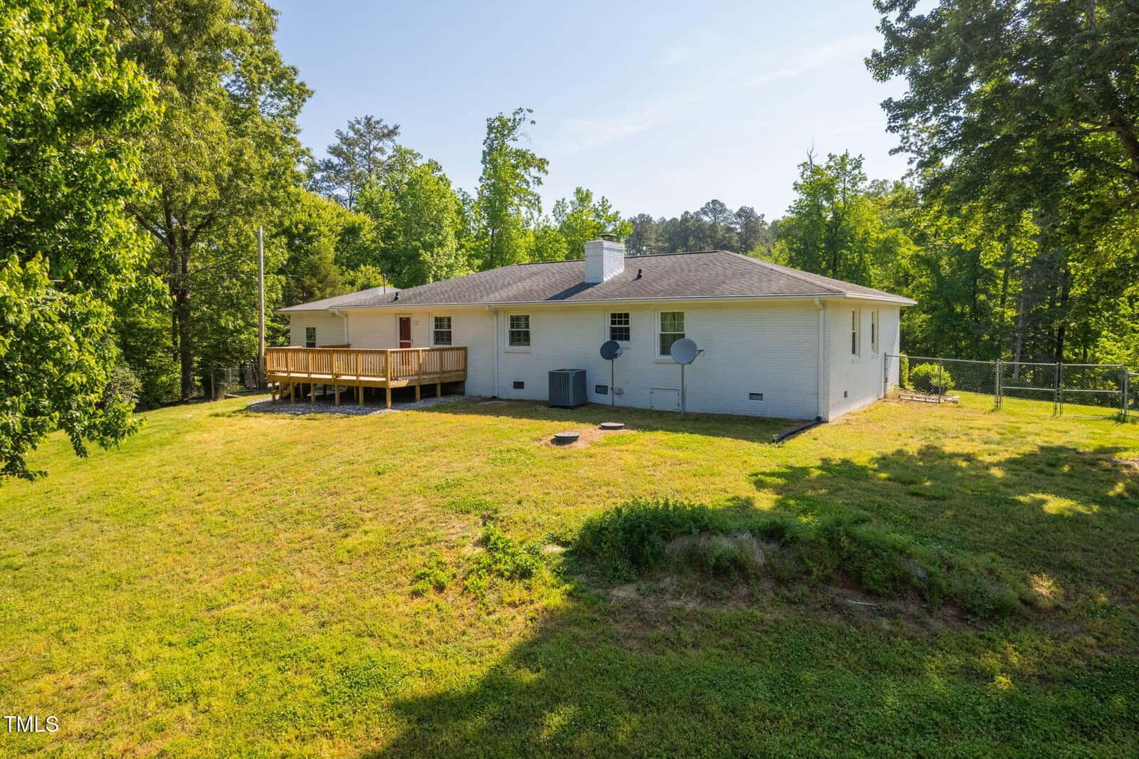 A single-story white brick house with a wooden deck on the back, two satellite dishes, and a large grassy backyard surrounded by trees under a clear sky.
