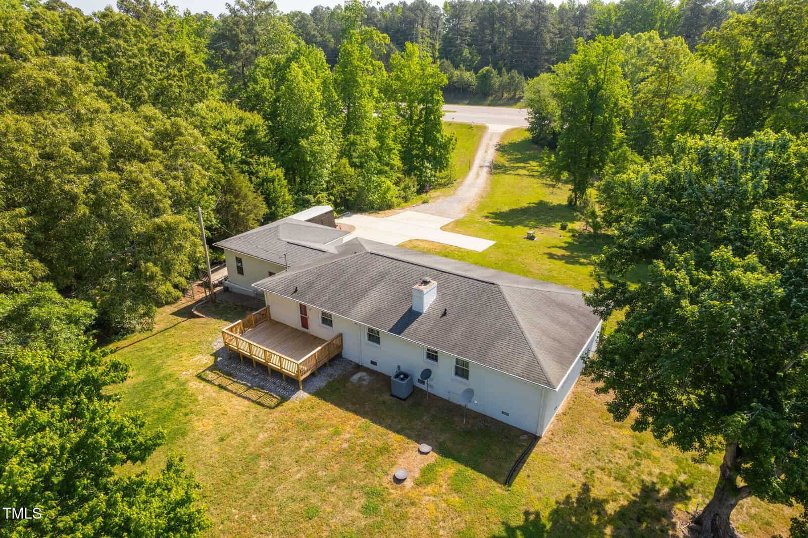 Aerial view of a single-story white house with a black roof, wooden deck, and large backyard surrounded by trees. A paved driveway leads to a road, and the area is bordered by greenery and forest.