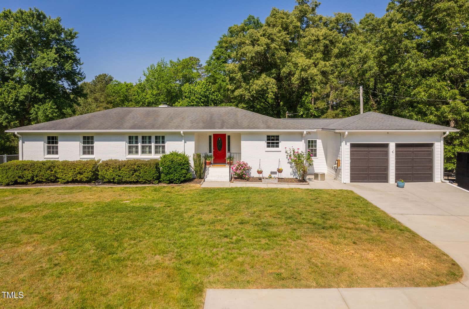 Charming single-story white brick home at 4200 US 1 Highway South, Kittrell, with a red front door, attached two-car garage with gray doors, manicured bushes, a spacious front yard, concrete driveway, and tall trees in the background.
