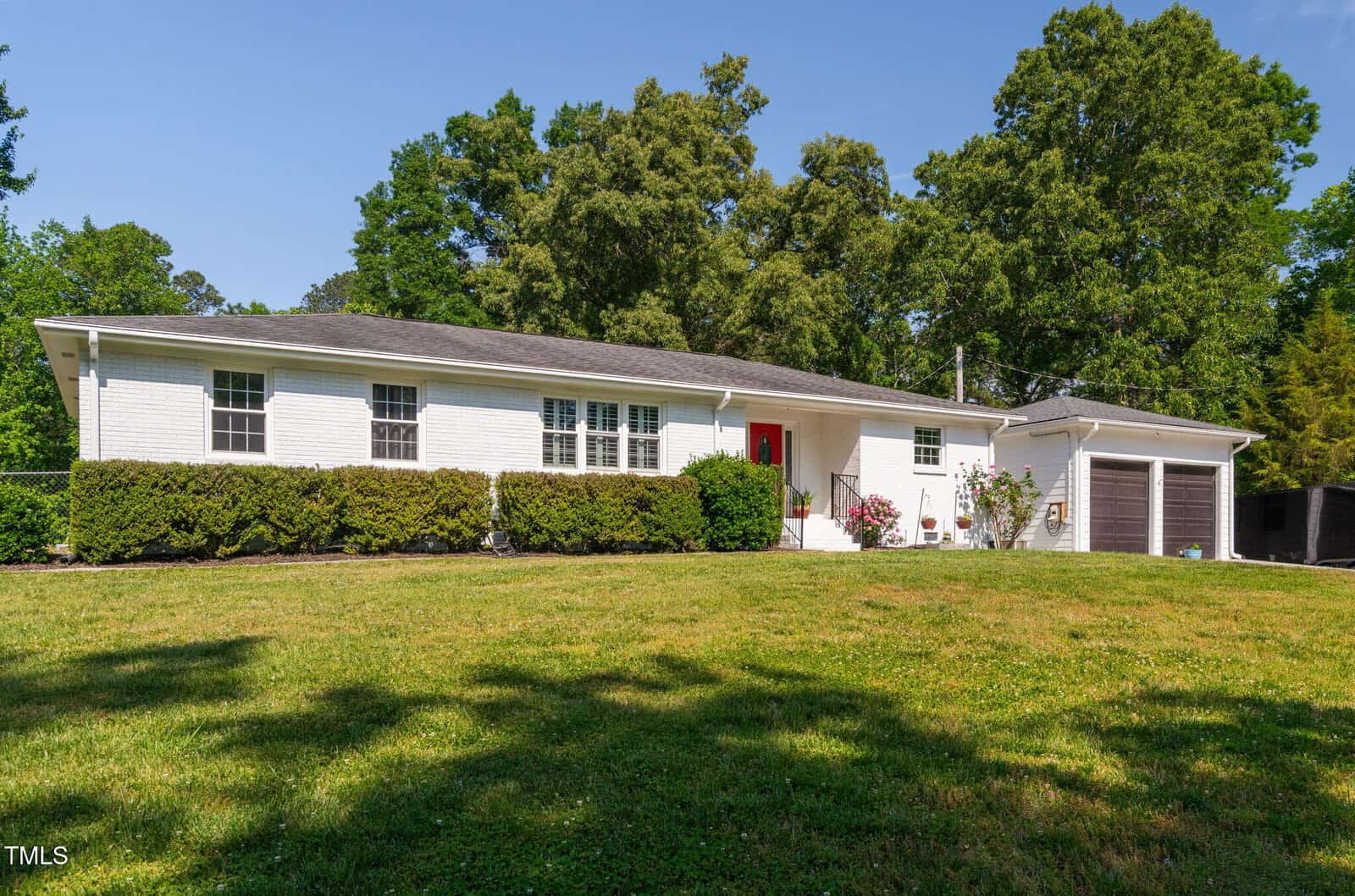 A single-story white brick house with a dark roof, red front door, attached double garage, trimmed hedges, and a large green lawn, surrounded by tall trees under a clear blue sky.