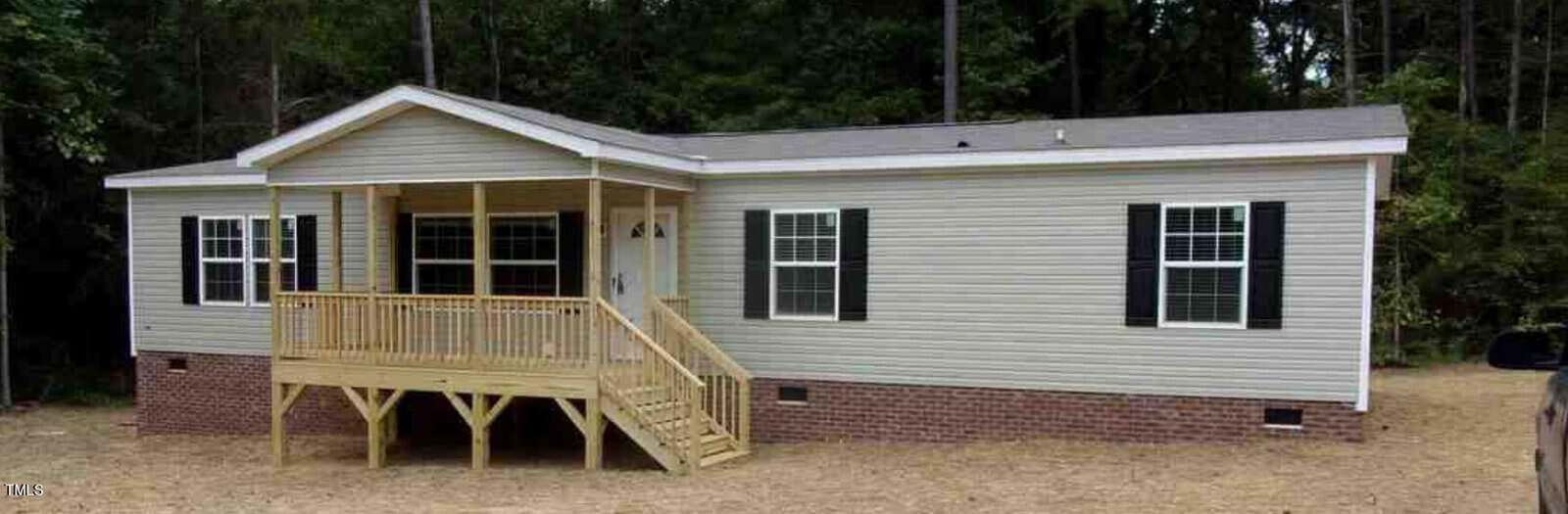 Located at 6646 Woodlawn Drive in Oxford, this beige manufactured home with a gray roof nestles among trees. It boasts a wooden porch with stairs leading to the entrance, complemented by white-framed windows and black shutters. The brick foundation and straw-covered yard complete the cozy setting.