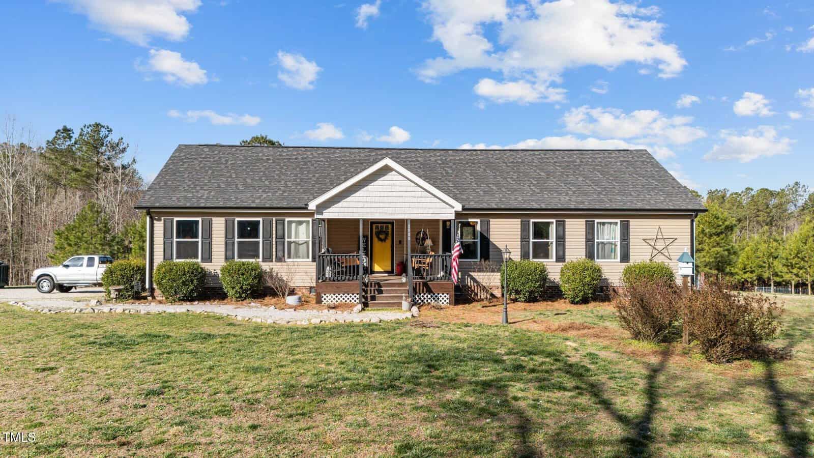 A single-story house with beige siding, dark shutters, and a front porch. An American flag hangs by the entrance, and a white pickup truck is parked to the left. The yard is grassy with shrubs and trees in the background.