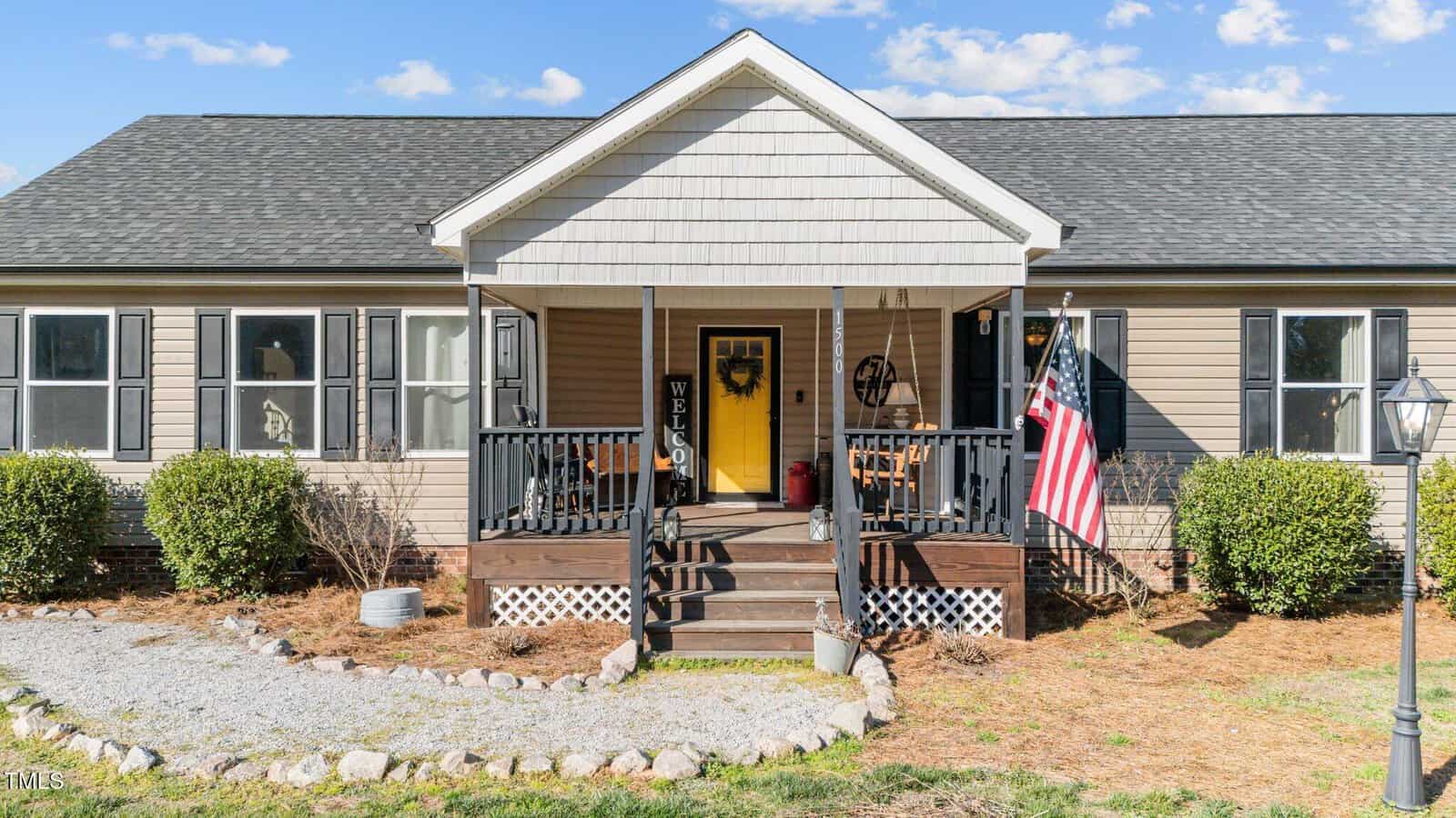 Single-story beige house with black shutters, a covered front porch, black railing, a yellow door, a “Welcome” sign, an American flag, and a gravel walkway bordered by rocks. Shrubs line the front of the house.
