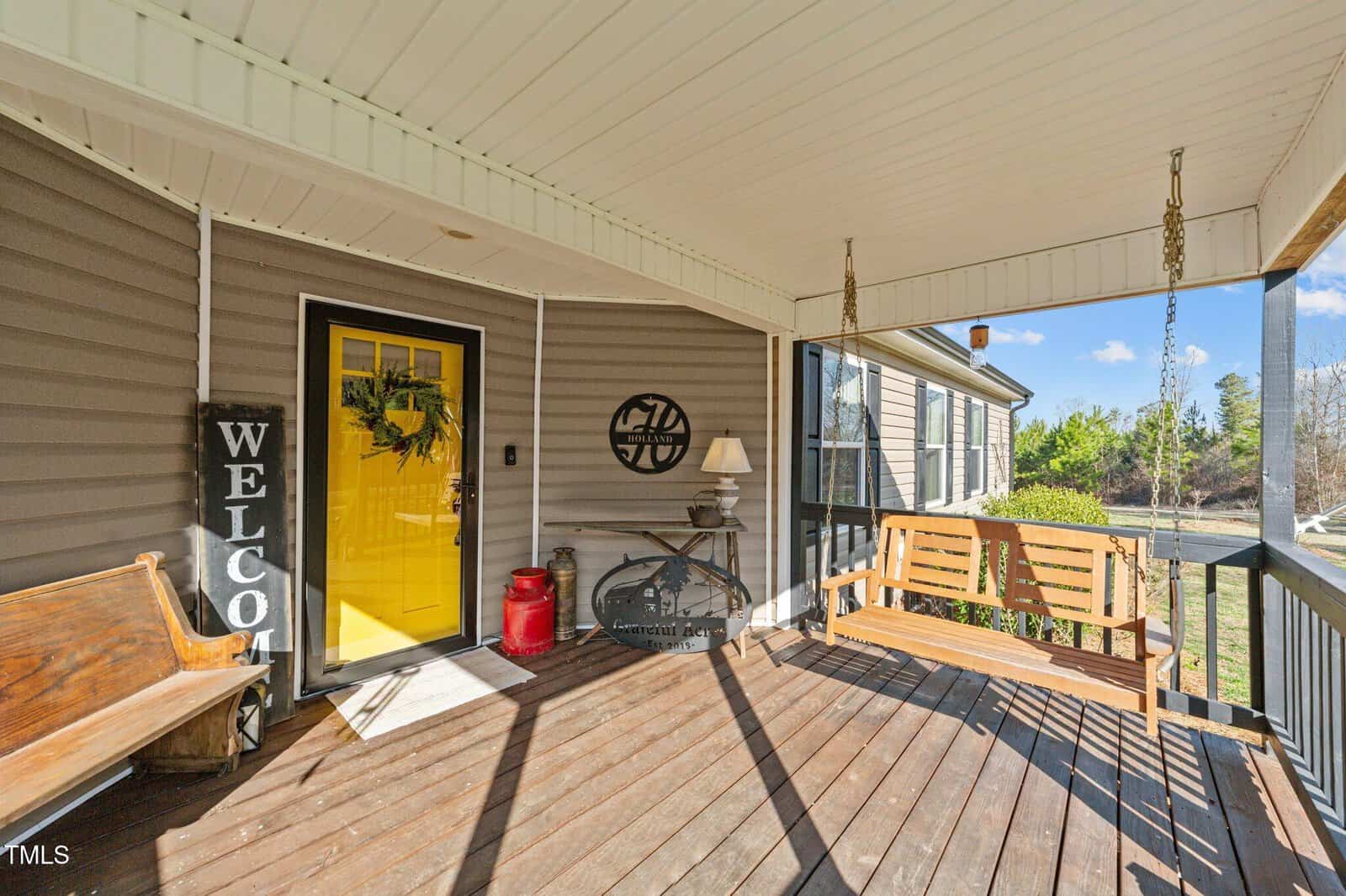 A covered porch with wooden flooring, a yellow front door, a welcome sign, a bench, a porch swing, a small table with a lamp, and decorative items, with sunlight streaming in.