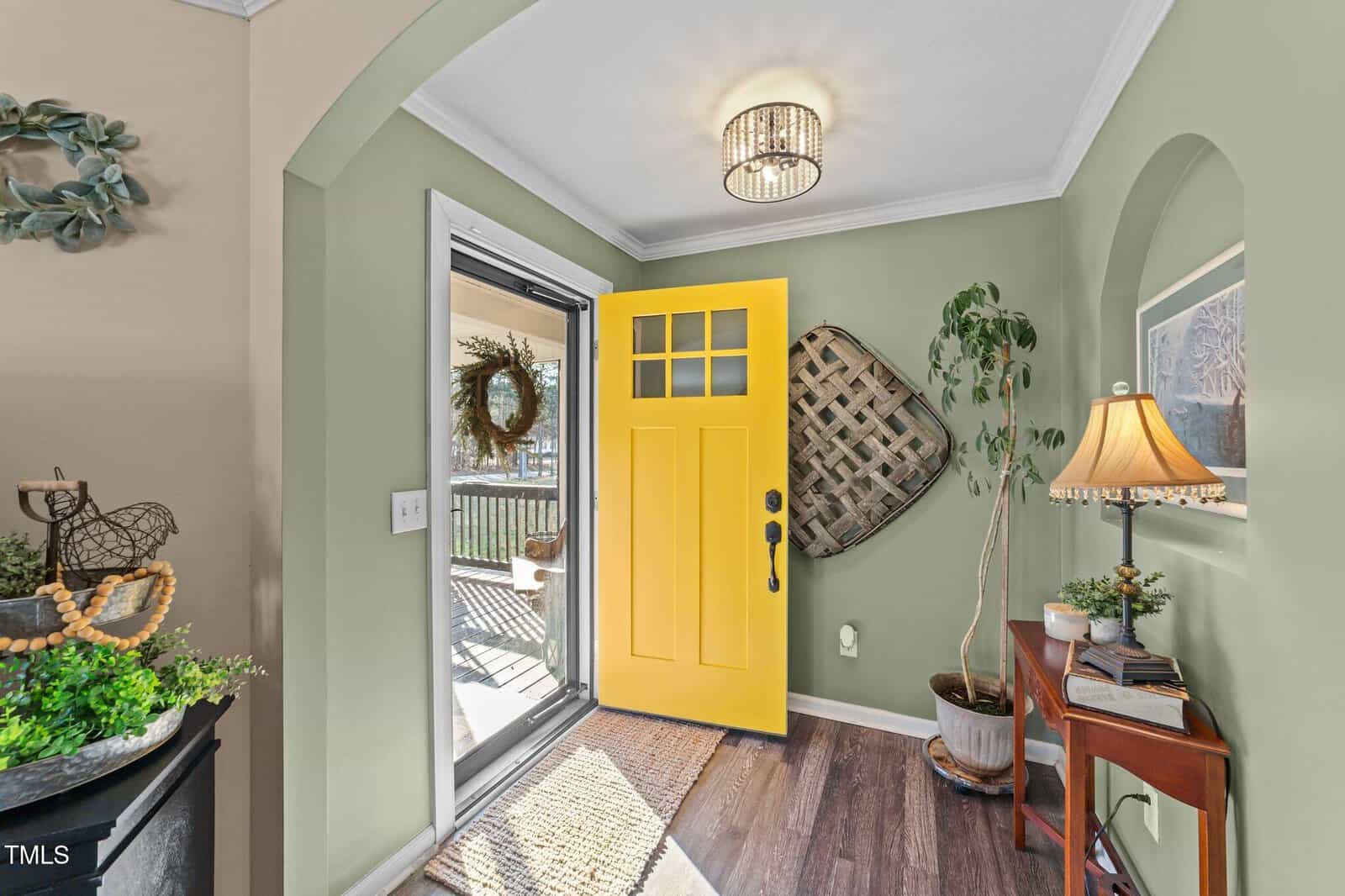 A small entryway features a bright yellow front door, green walls, wood flooring, a round ceiling light, a table lamp on a wooden side table, houseplants, and various wall decorations. Sunlight enters from the open door.