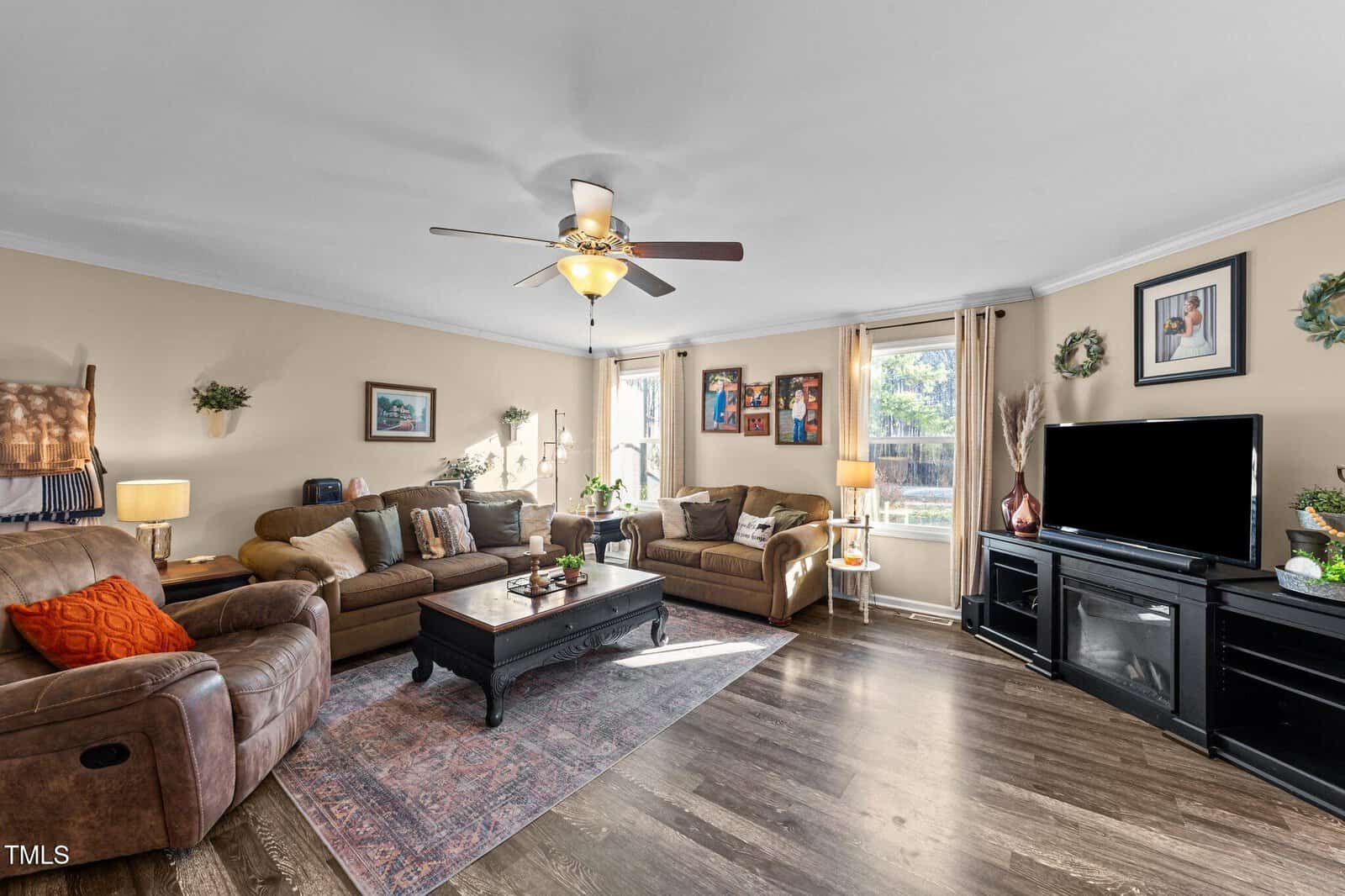 A living room with two brown sofas, a recliner, a coffee table, a TV on a stand, wall art, and potted plants. Sunlight comes through large windows with sheer curtains, illuminating the wood floor.