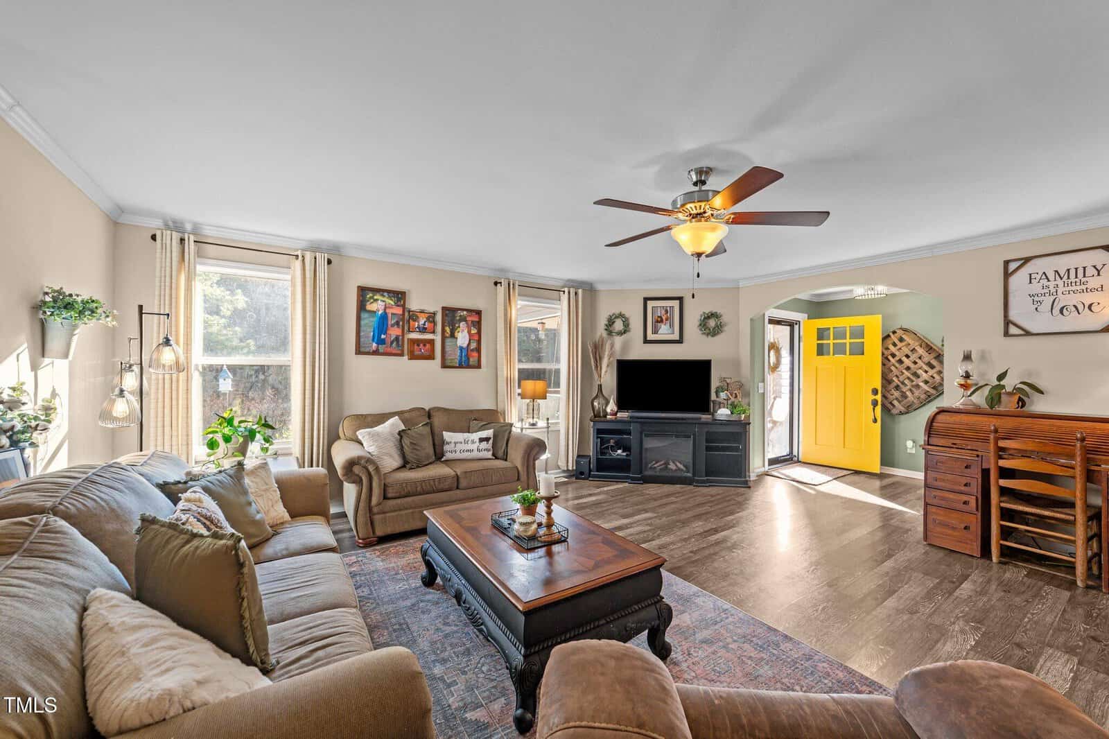Living room with two sofas, a coffee table, TV on a stand, a ceiling fan, a yellow front door, wall art, indoor plants, and wooden floors. Sunlight enters through two windows with beige curtains on the left wall.