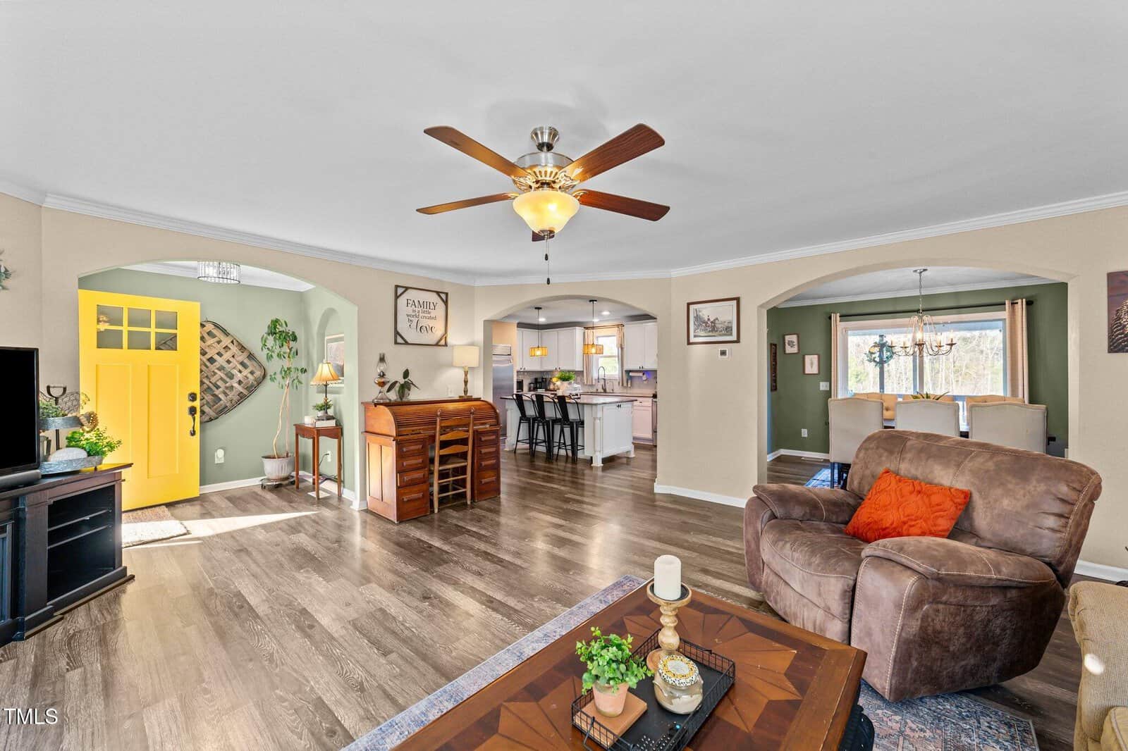 Living room with brown flooring, ceiling fan, recliner, and coffee table. View of a kitchen with white cabinets, dining area, and entry with a yellow door and green accent wall. Natural light fills the space.