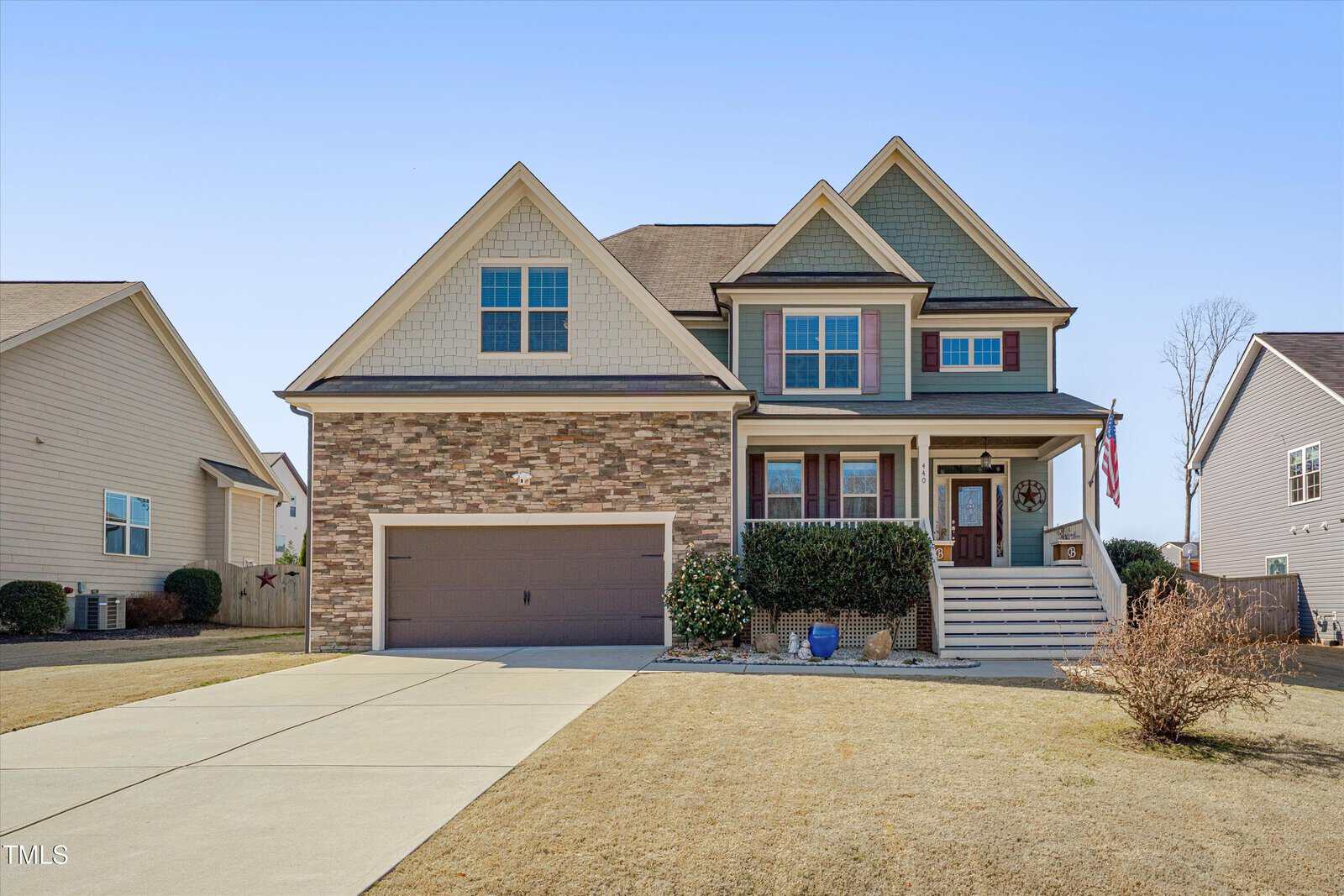 A two-story suburban house with a brick and siding exterior, double garage, covered front porch with steps, and a manicured lawn. The driveway leads up to the garage, and neighboring houses are visible on both sides.