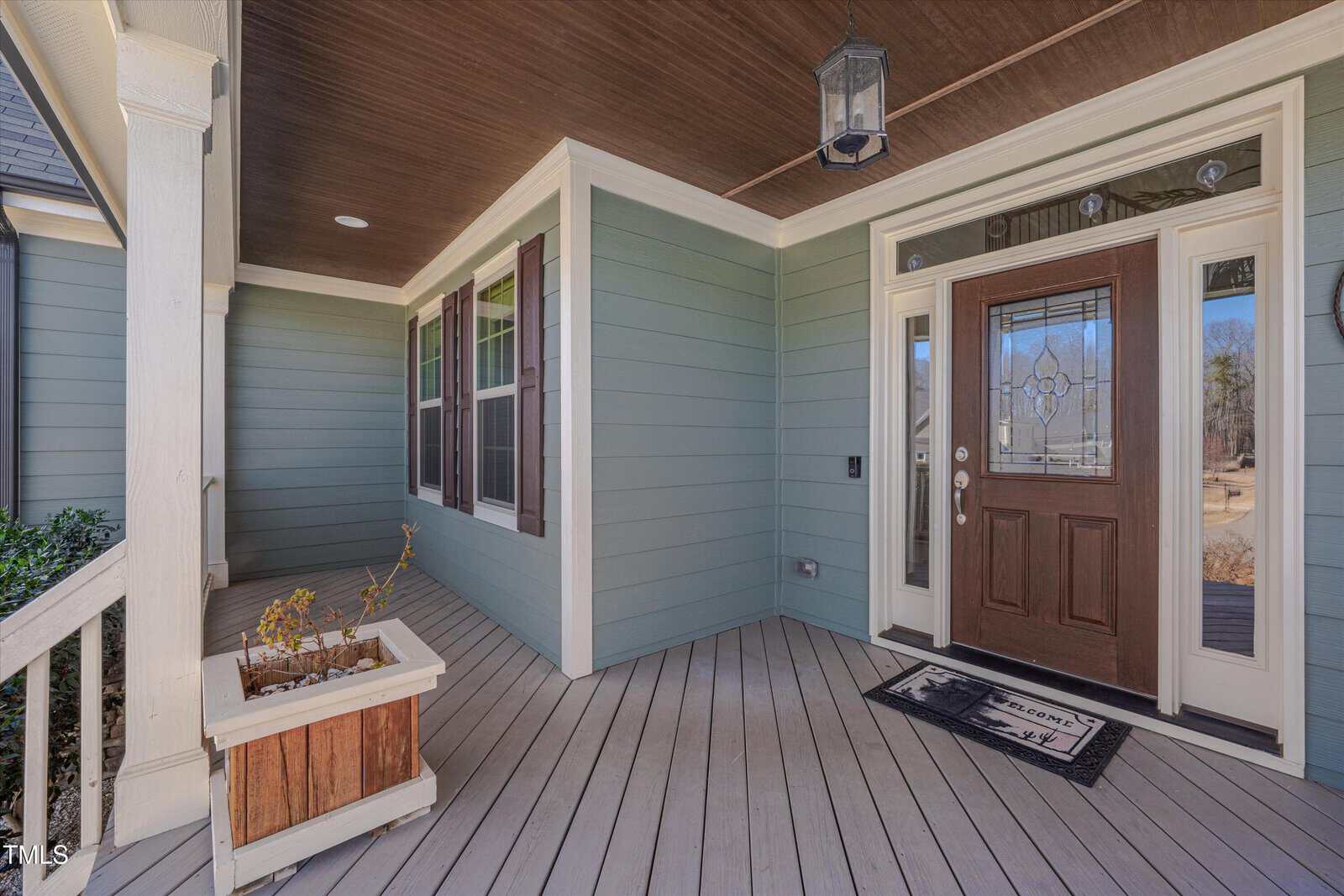 A covered front porch with light green siding, white trim, and brown accents features a wooden planter with dried plants, a brown front door with decorative glass, sidelights, a doormat, and a hanging lantern.
