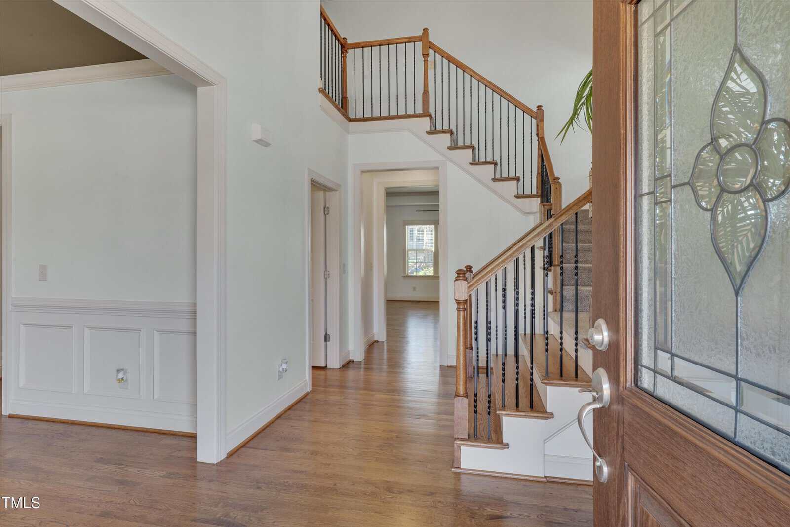 View of a home's entryway featuring hardwood floors, white walls with wainscoting, an open staircase with wooden handrails and black balusters, and a partially open wooden door with decorative glass.