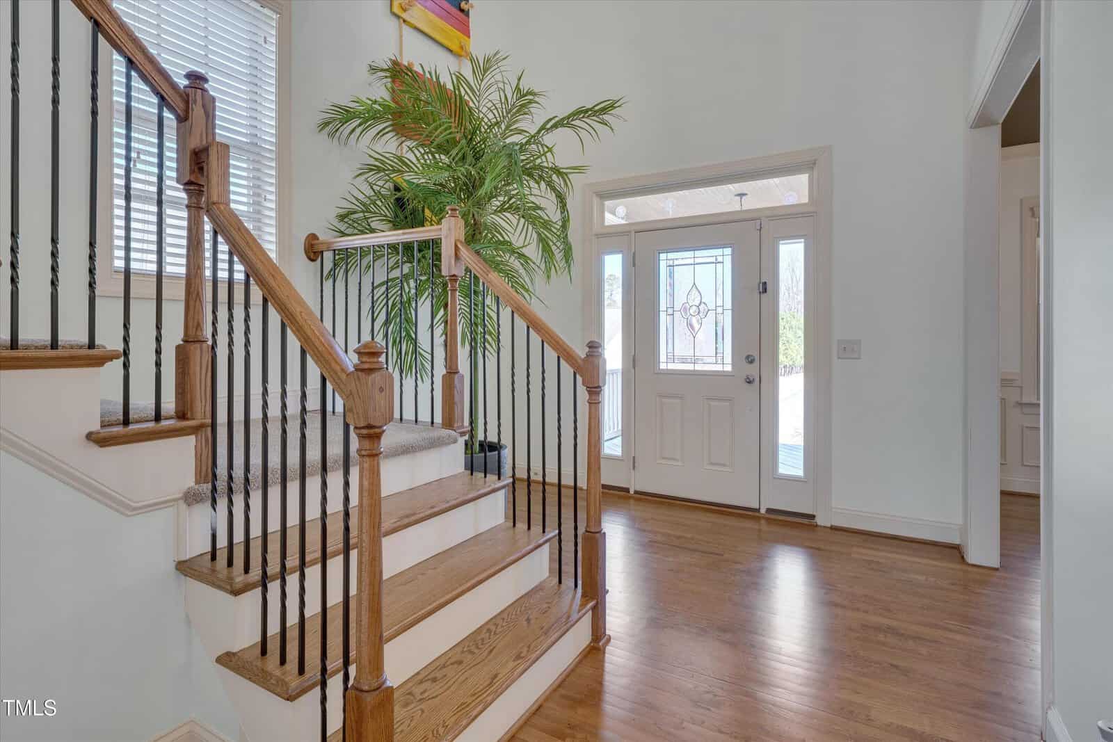 A wooden staircase with carpeted steps and black spindles is next to a front door with glass panels. The entryway has hardwood floors, a large potted plant, and a window with blinds.
