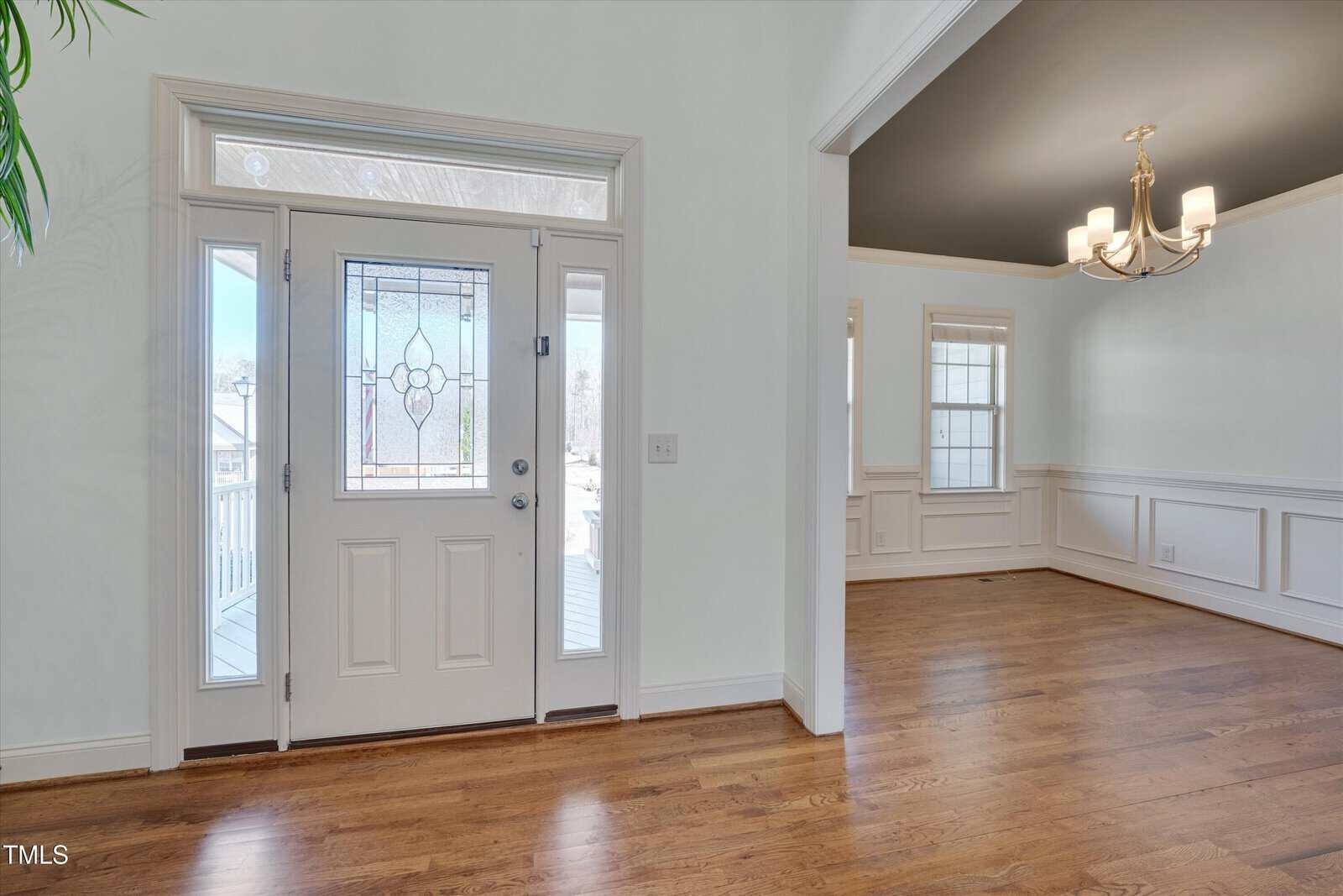 A view of a home's entryway with a white front door featuring decorative glass panels, adjacent to a dining room with hardwood floors, white wainscoting, and a ceiling-mounted chandelier.